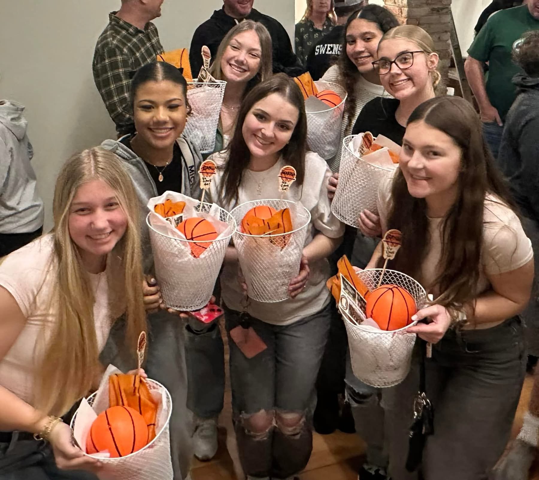 A group of young people at a celebration, holding baskets with small basketballs and Halloween-themed decorations, smiling and posing together indoors.