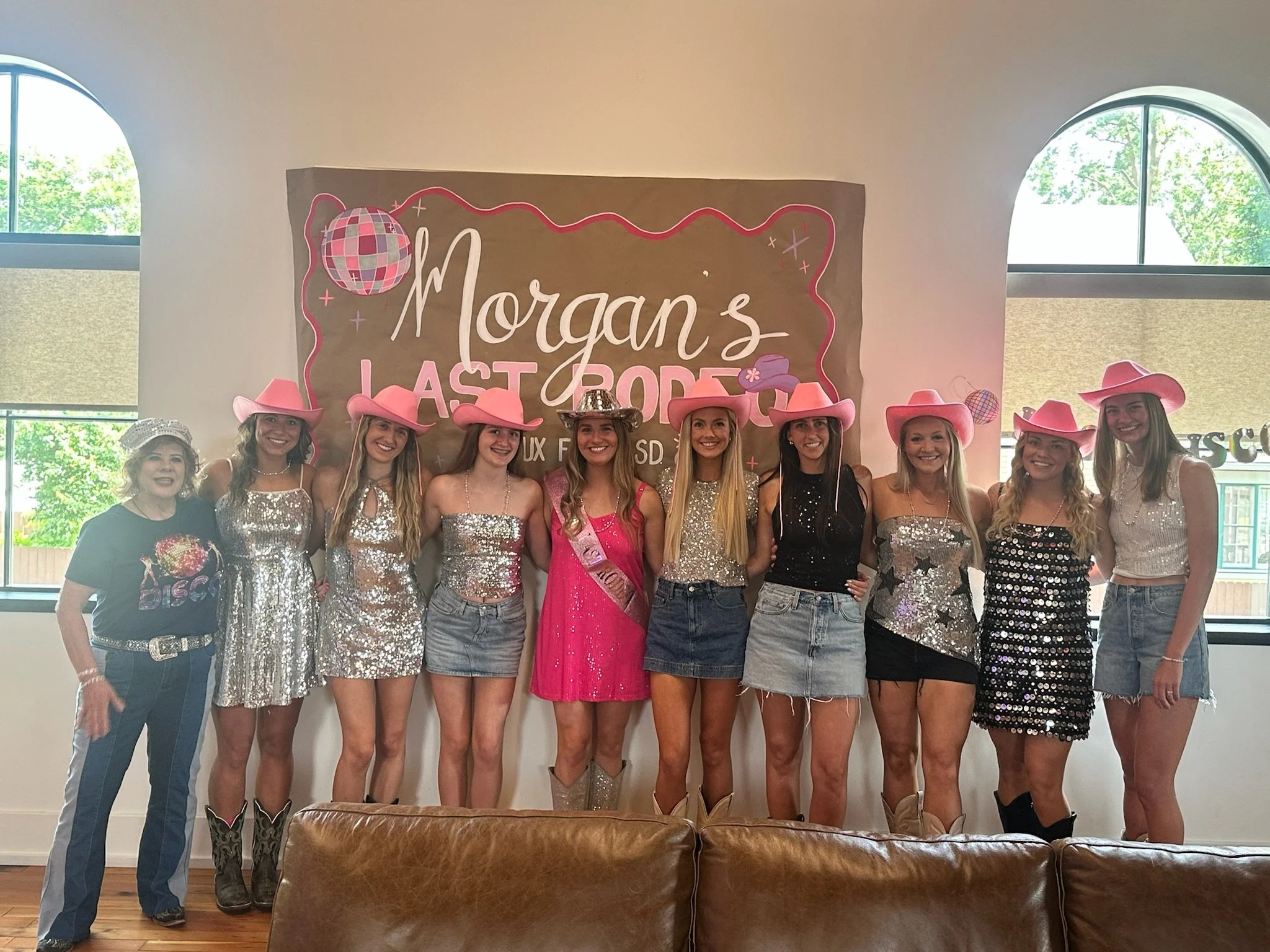 Group of women and girls dressed in western-inspired outfits and cowboy hats, standing in front of a banner that says 'Morgan's Last Rodeo' at a celebration or event.