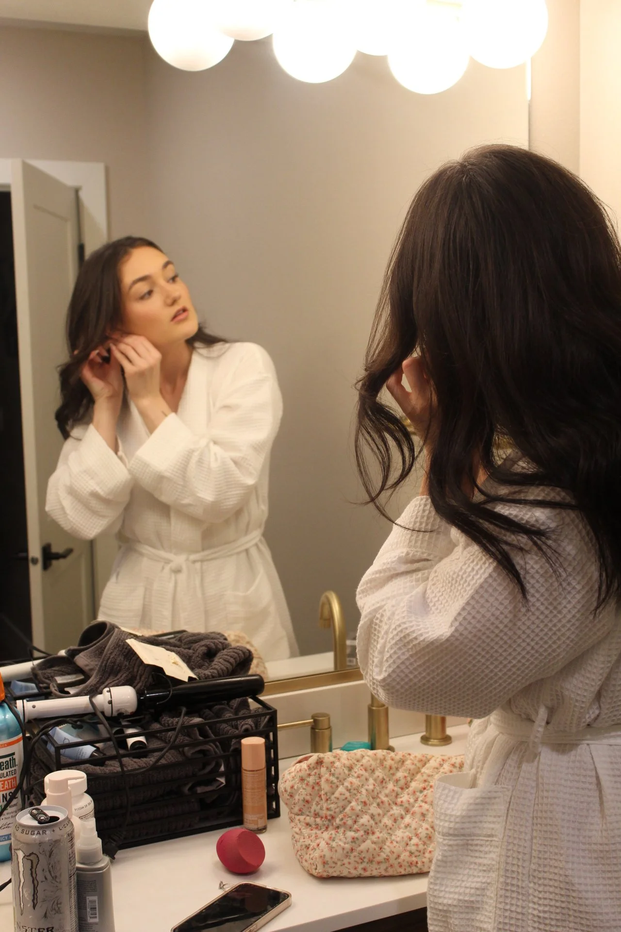 Young woman in a white robe looks at herself in the bathroom mirror while fixing her hair, with various hair and beauty products on the counter.