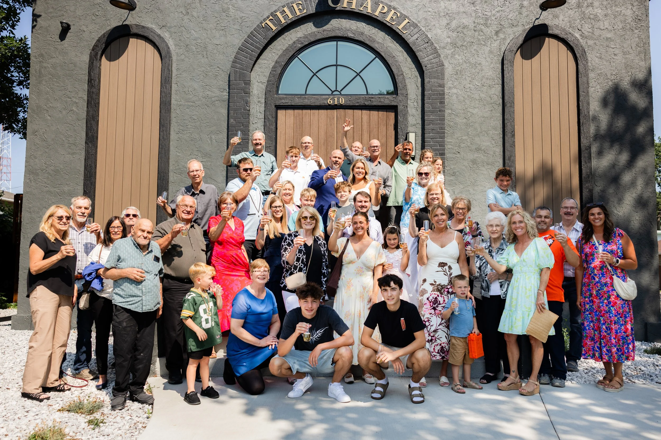 A large group of people celebrating in front of a church building, raising glasses in a toast. The group includes men, women, children, and elderly individuals, all smiling. The church has a sign with the name "The Chapel" and the number "610 above t