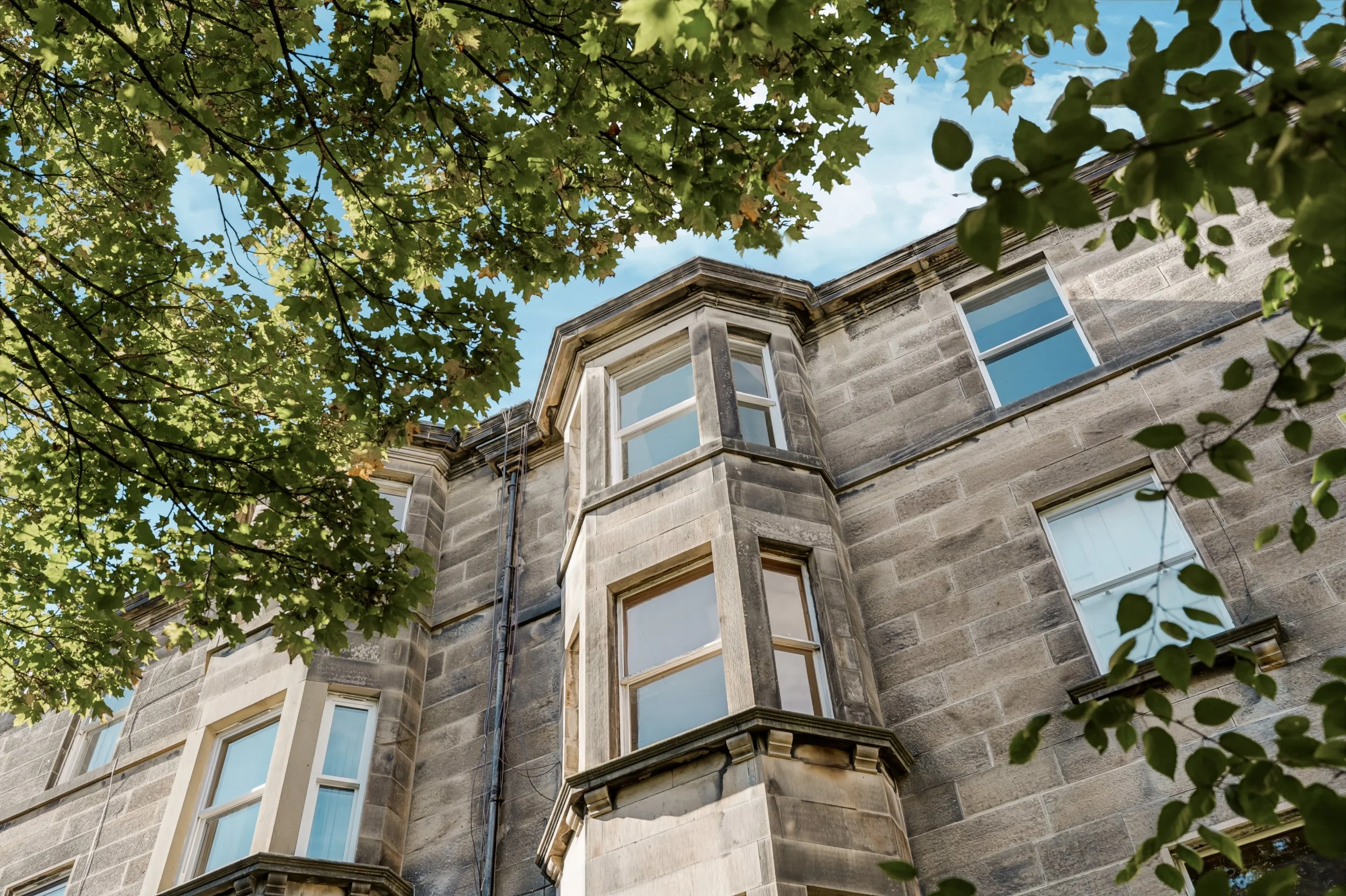 Looking up at a stone building with a bay window, partially obscured by green tree leaves against a blue sky with some clouds.
