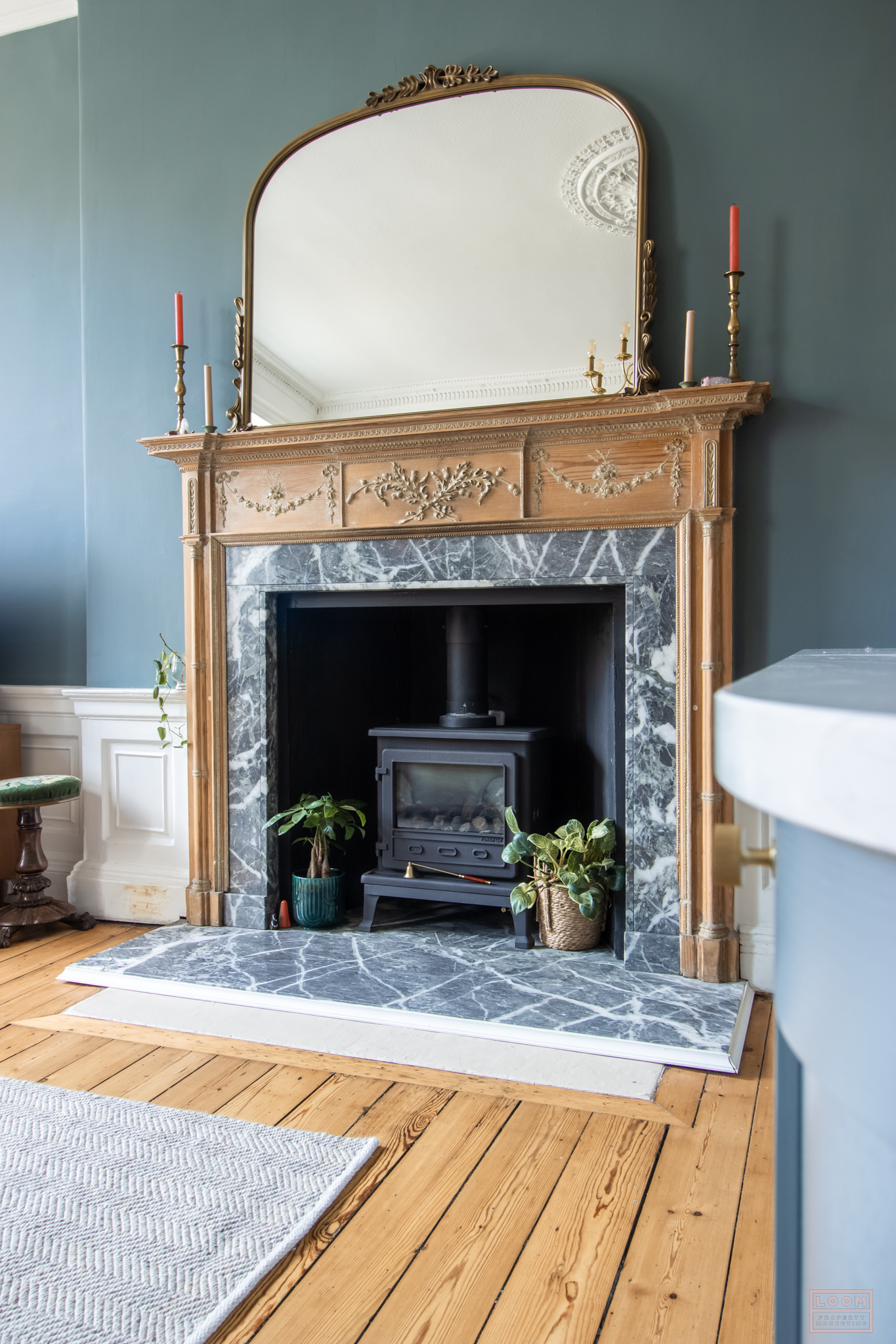 A fireplace with a carved wooden mantel, a black stove inside, potted plants on each side, and a large mirror with a decorative gold frame above. Candles are placed on the mantel, and part of a white rug is visible on a wooden floor.
