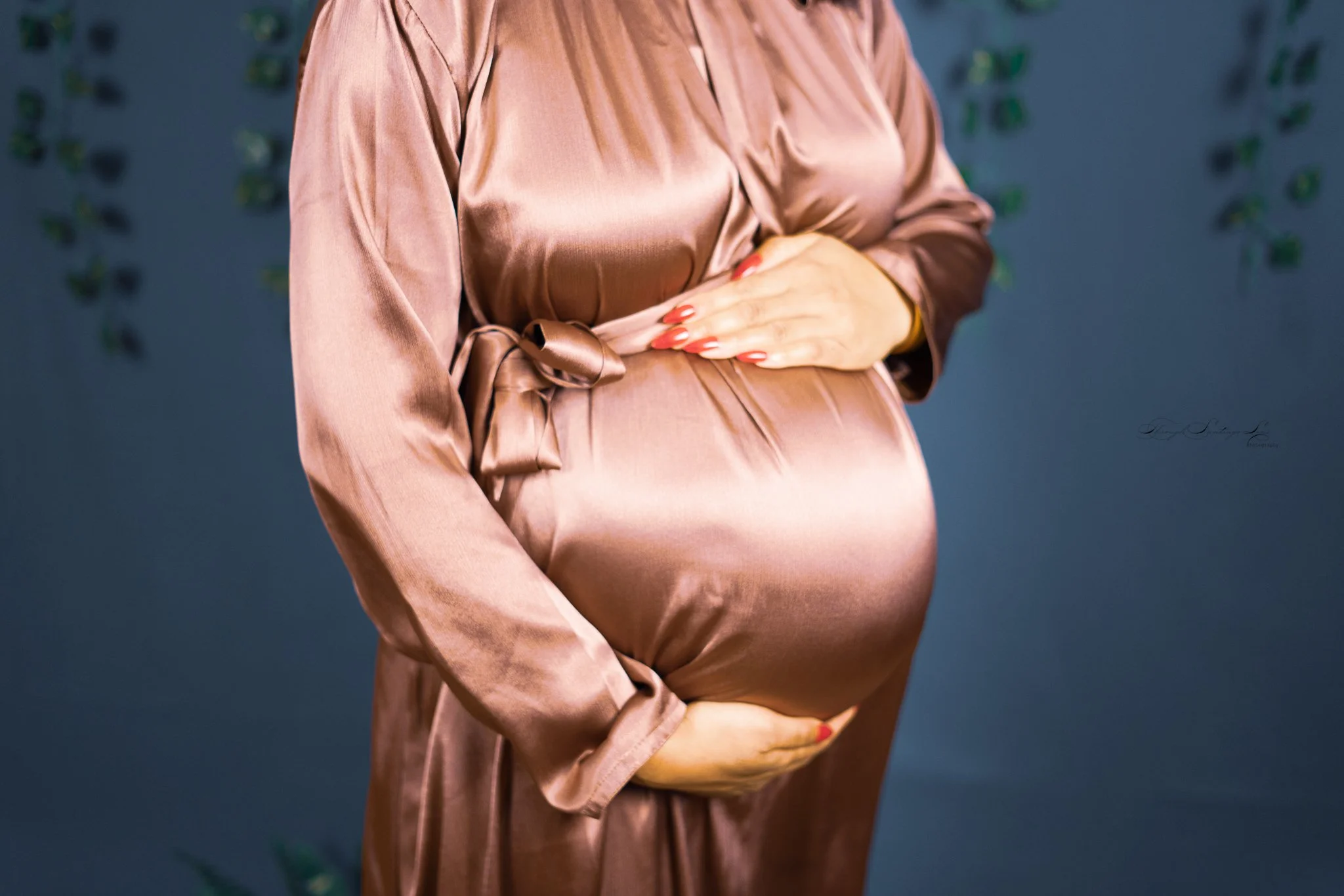 Close-up of a pregnant woman wearing a silky satin dress in a soft pink color, with her hand gently resting on her belly, standing against a dark, patterned background.