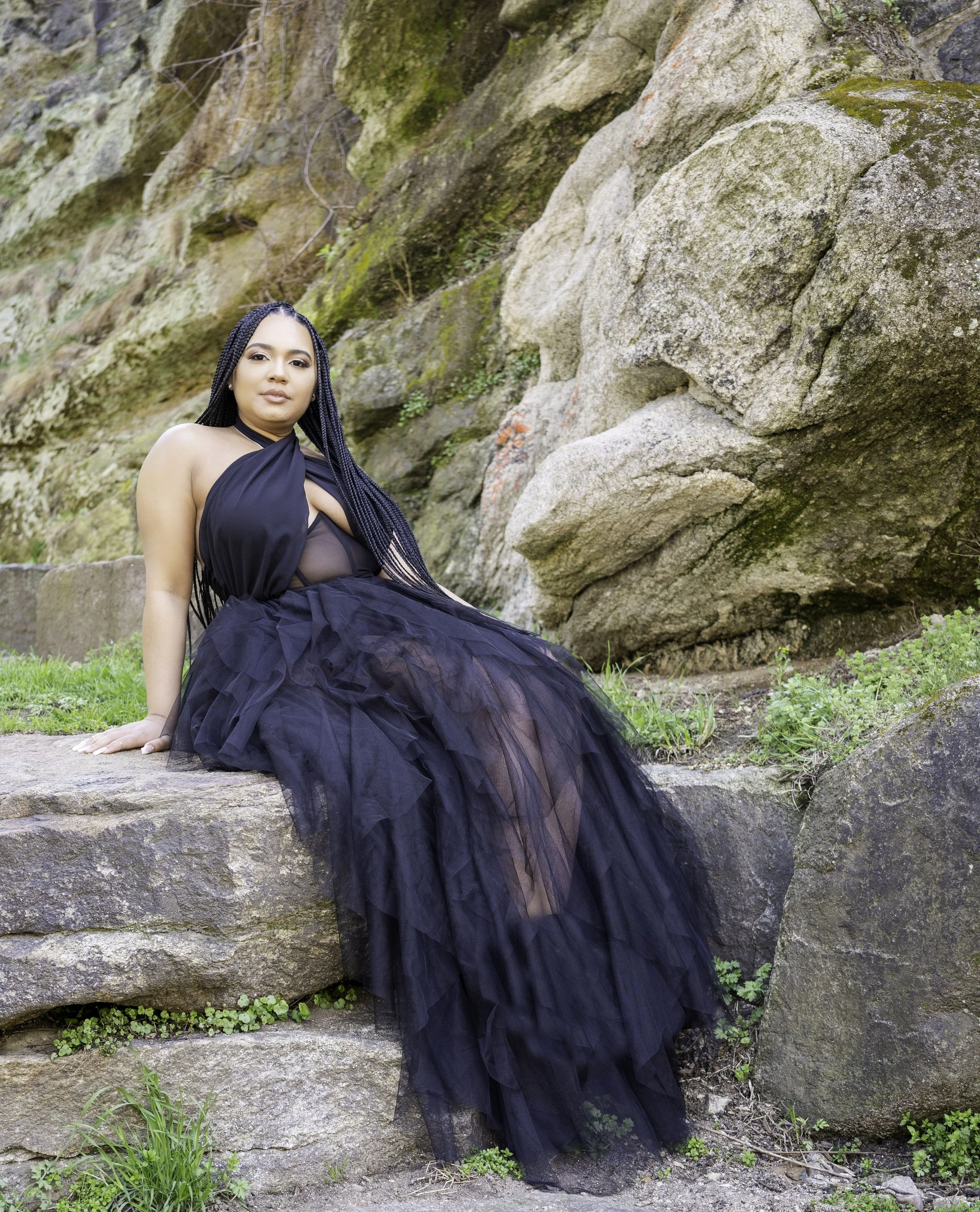 A woman with long braided hair sitting on a rock near a large rock formation, wearing a black strapless dress with a full tulle skirt, outdoors with grass and small plants surrounding her.