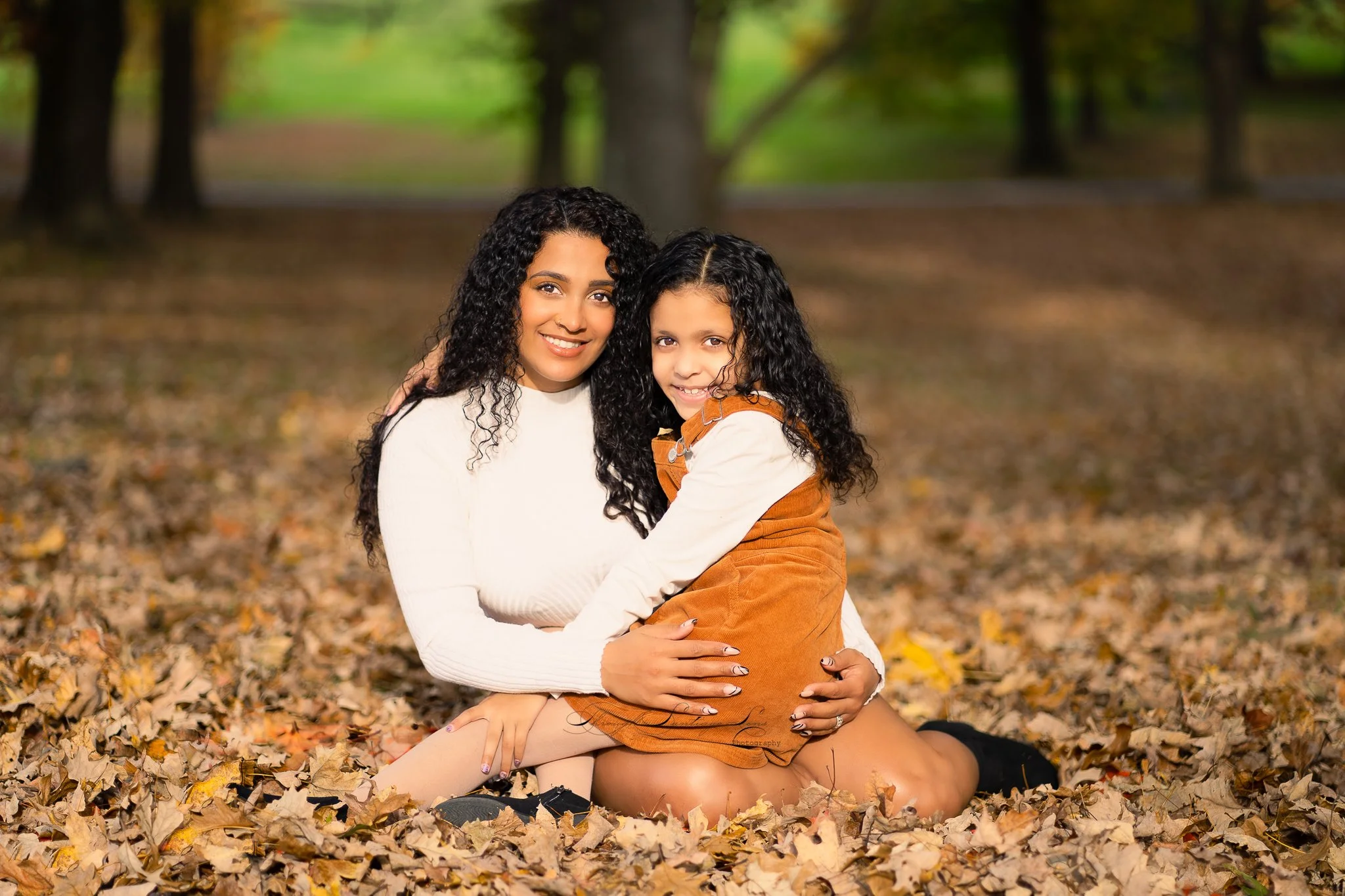 A woman and a girl with curly hair sitting on fallen autumn leaves in a wooded park, hugging and smiling at the camera.