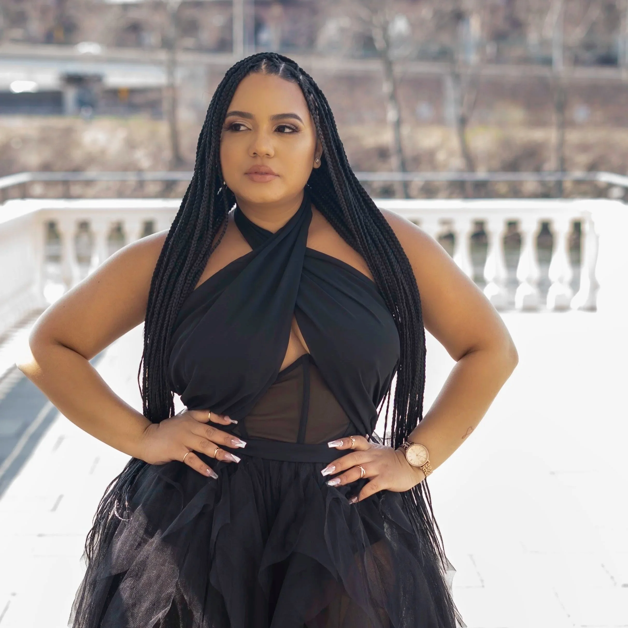 A woman with long braided hair standing outdoors on a balcony with a river and trees in the background, wearing a black dress with a halter neckline, and jewelry including earrings, rings, and a wristwatch.
