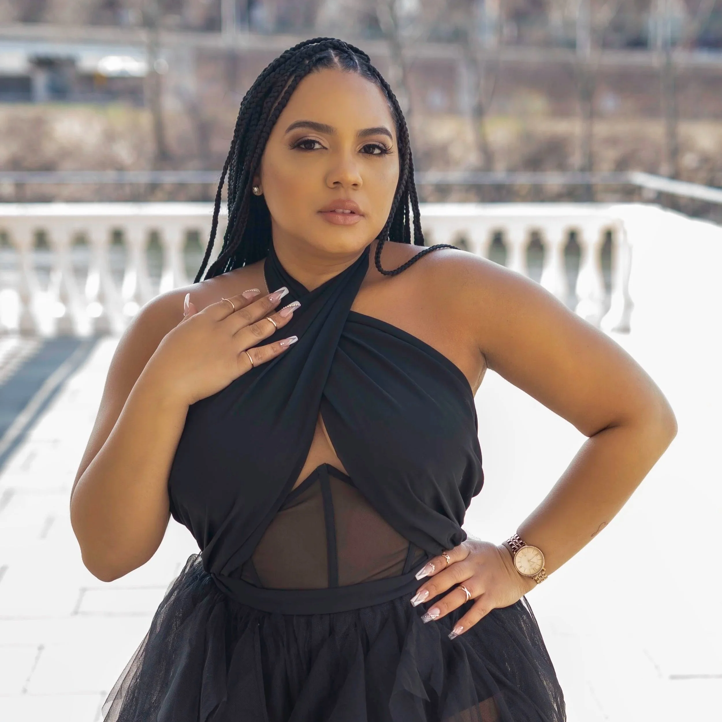A woman with long braided hair dressed in a black halter top, wearing rings and a gold watch, standing outdoors near a white railing with trees in the background.