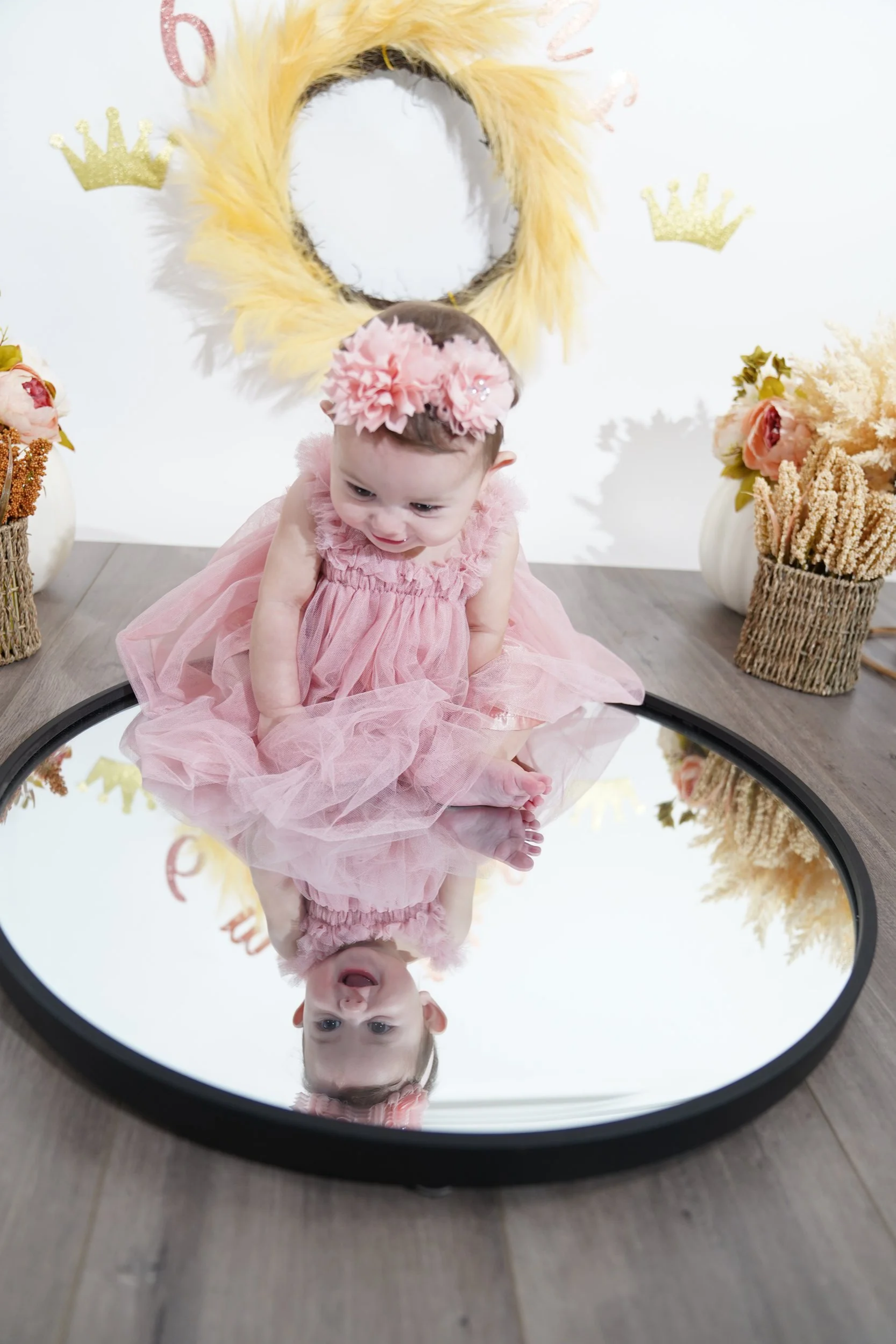 Young girl in a pink dress and floral headband looking at her reflection in a round mirror on the floor, with a fluffy yellow mustache and crown decorations in the background.