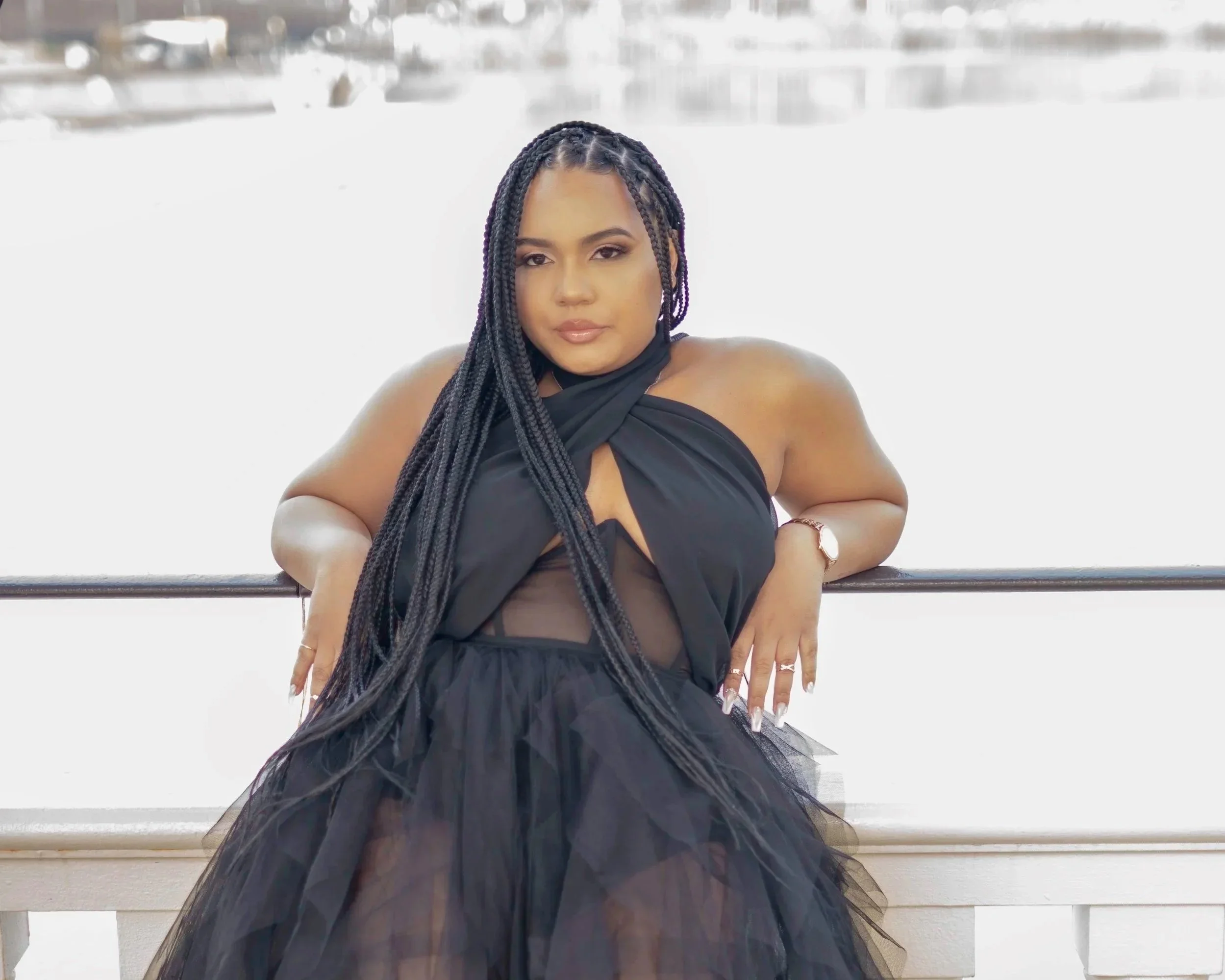 A woman with braided hair wearing a black dress with sheer details, leaning on a railing with her arms crossed in front of a background of a lake and a cloudy sky.