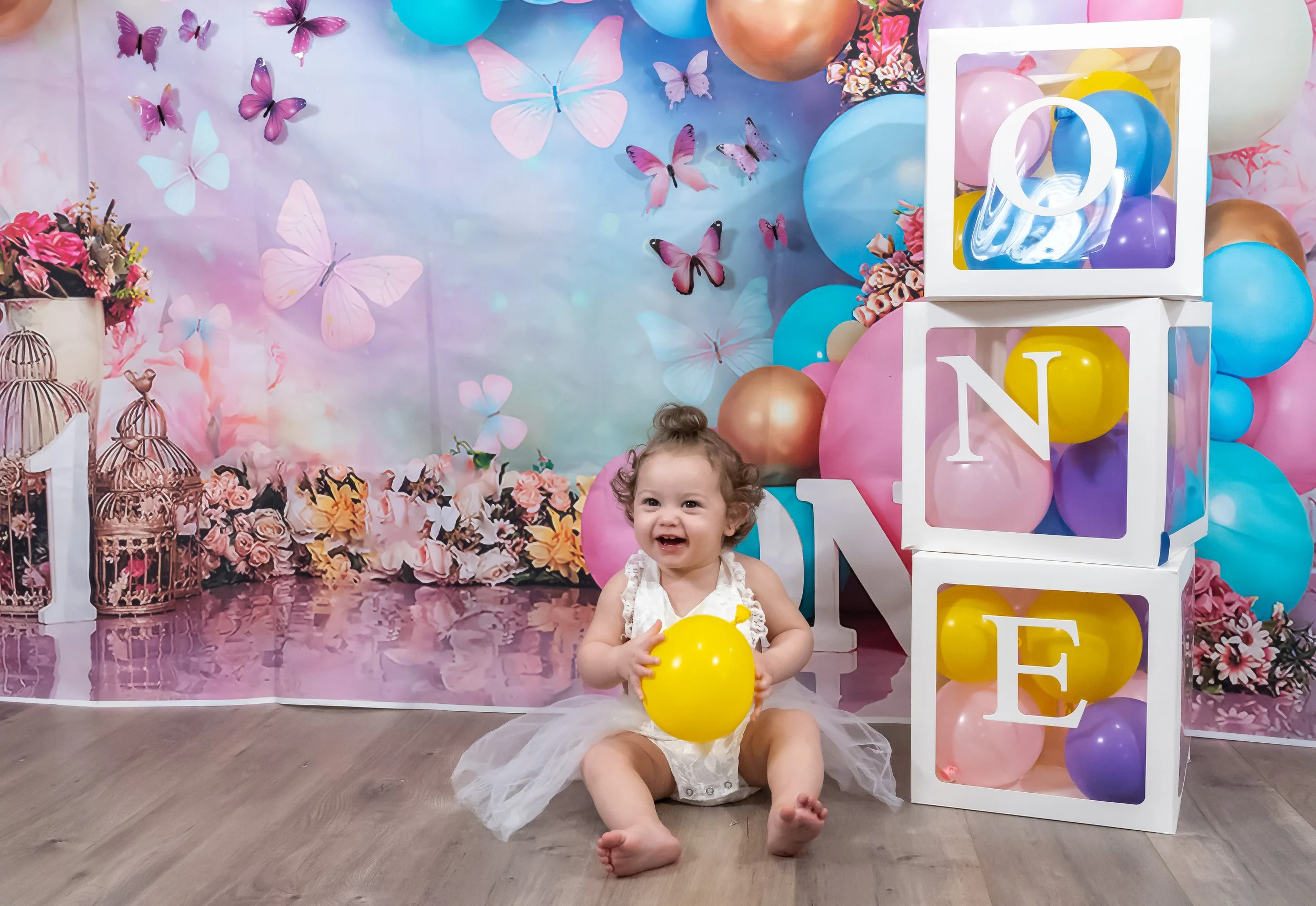 A smiling young girl sitting on the floor holding a yellow balloon, with a colorful butterfly-themed backdrop and balloon decorations behind her.