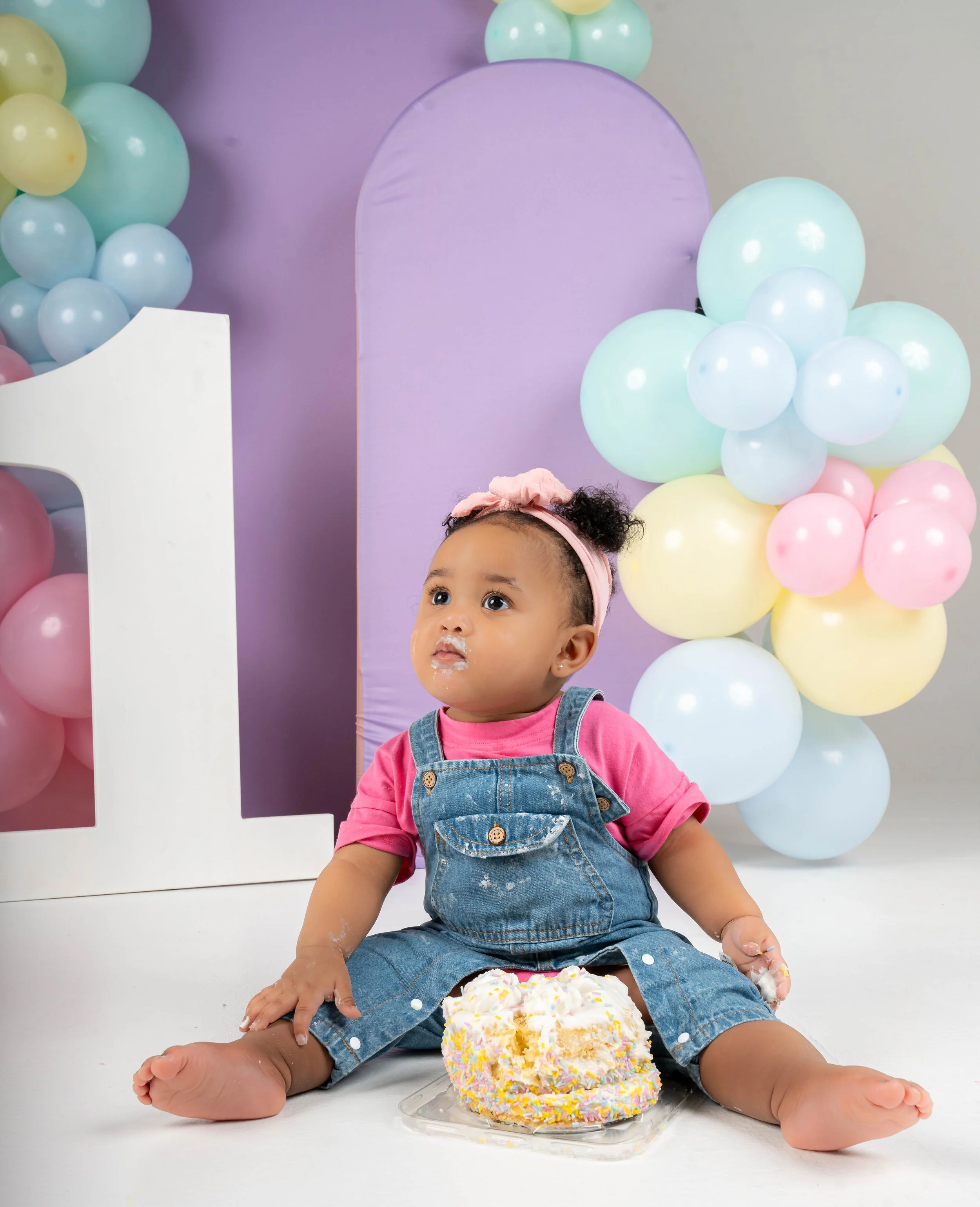 A young girl with curly hair, wearing a pink shirt, denim overalls, and a pink headband, sitting on the floor in front of a large birthday cake with white frosting and colorful sprinkles, surrounded by pastel-colored balloons against a backdrop of pu