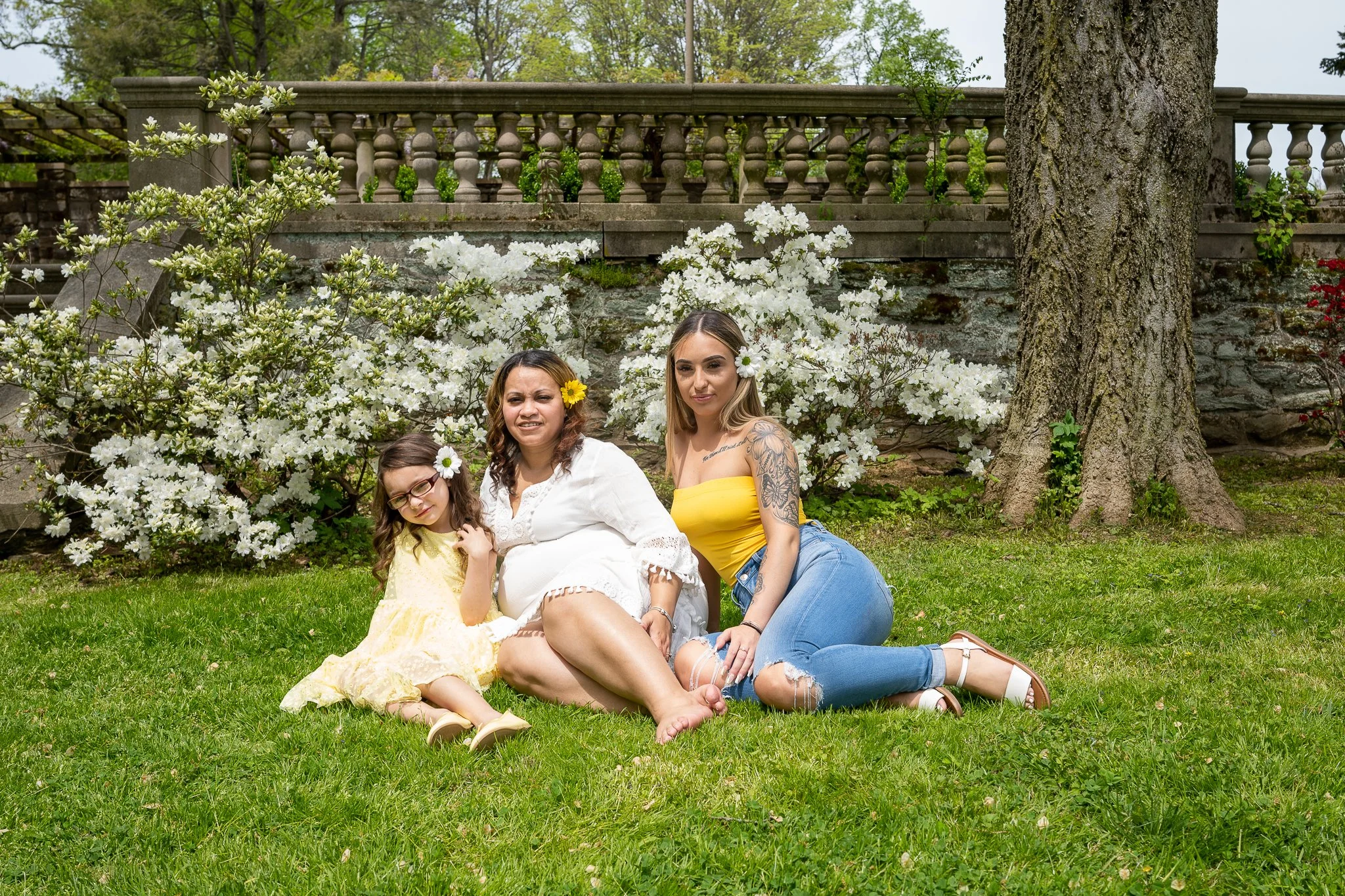 A group of four women and a young girl sitting and lying on grass in a garden with blooming white flowers, trees, and a stone border with a decorative stone balustrade in the background.