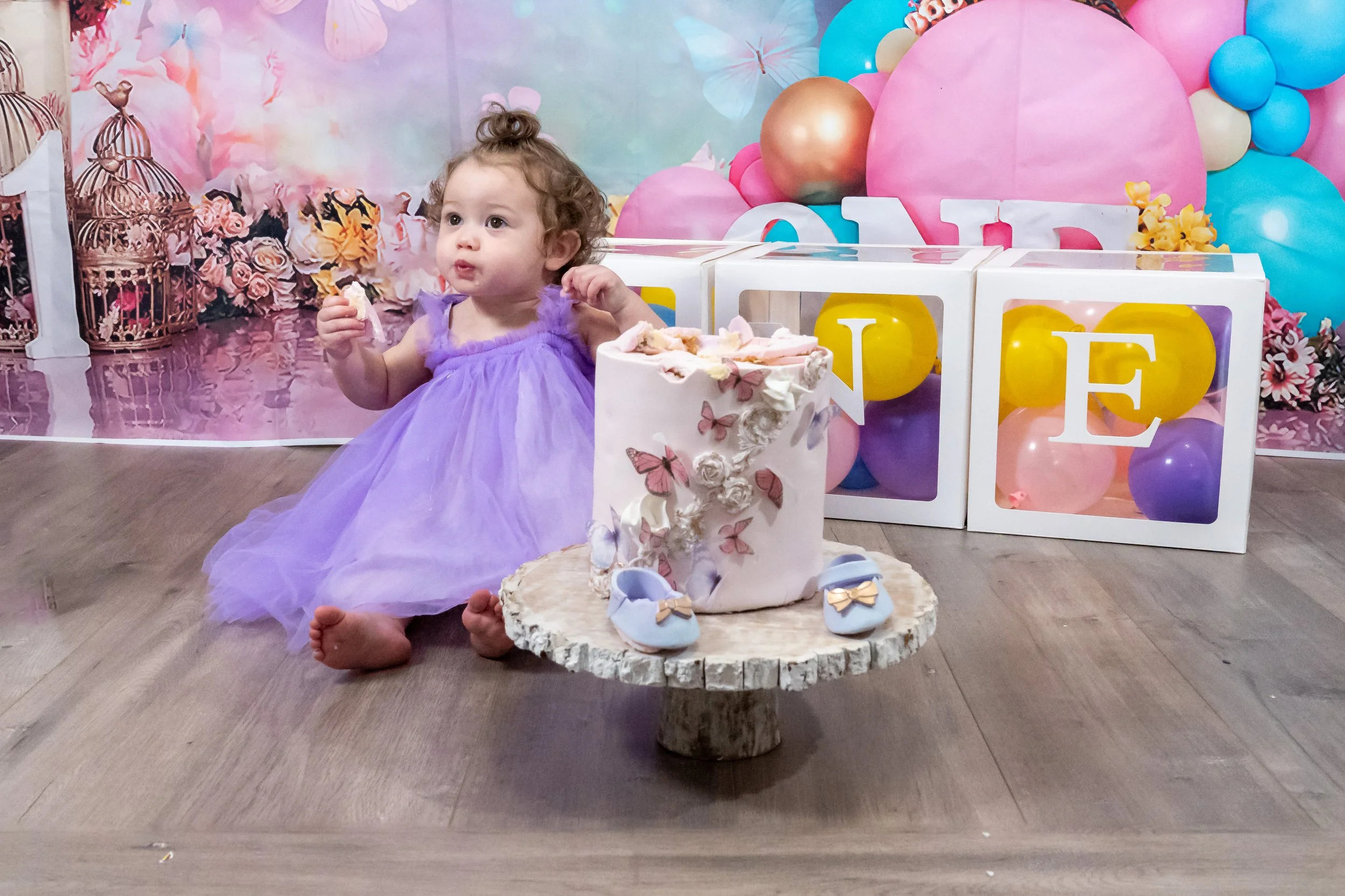 A young girl in a purple dress sitting on the floor at a birthday party, with a large pink and blue balloon backdrop, a cake decorated with butterflies and flowers in front of her, and colorful letter blocks spelling 'LOVE' behind her.