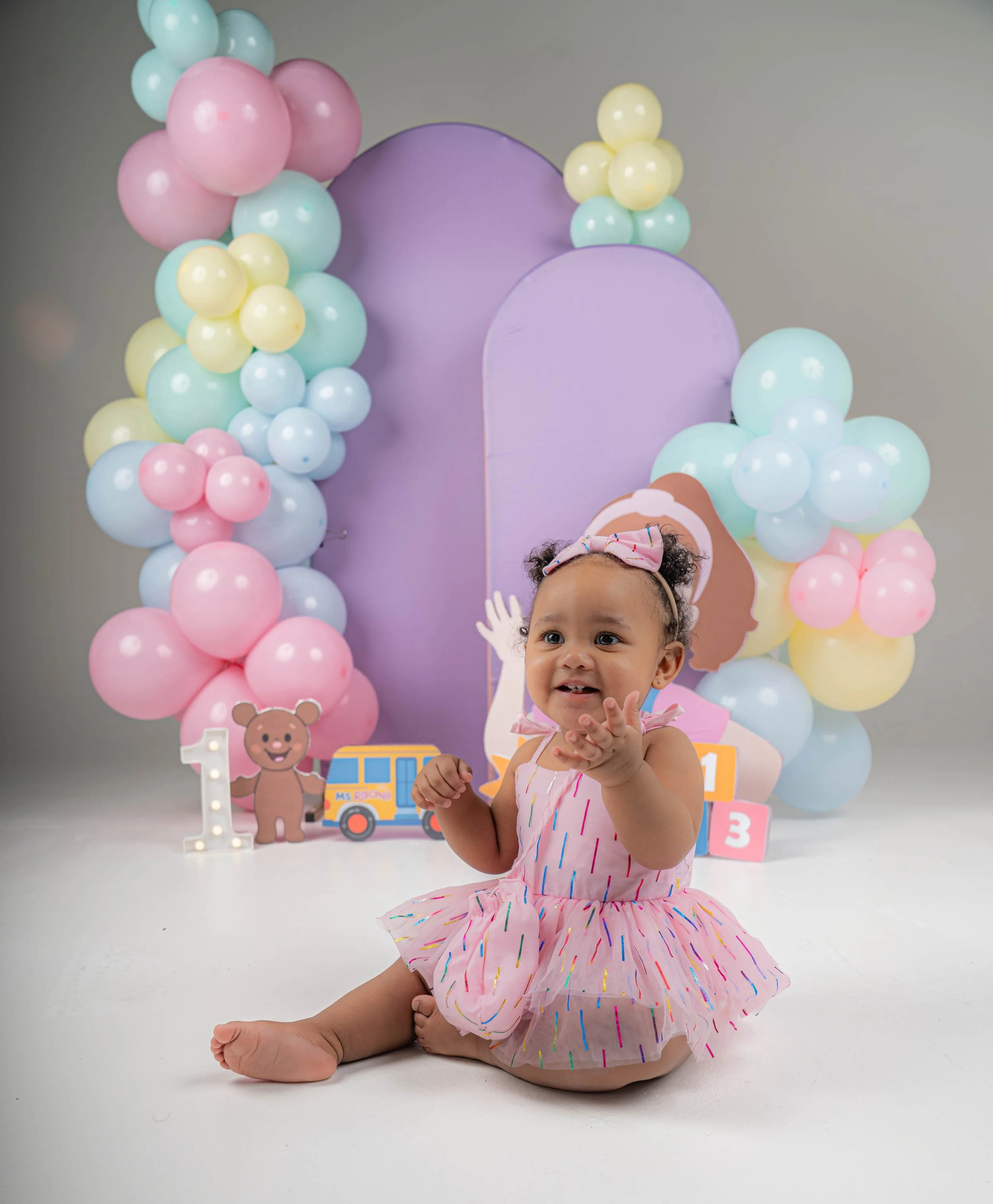 Young girl with curly hair, wearing a pink dress with colorful sprinkles, sitting on the floor in front of a pastel-colored birthday display with balloons, cake, and decorations.
