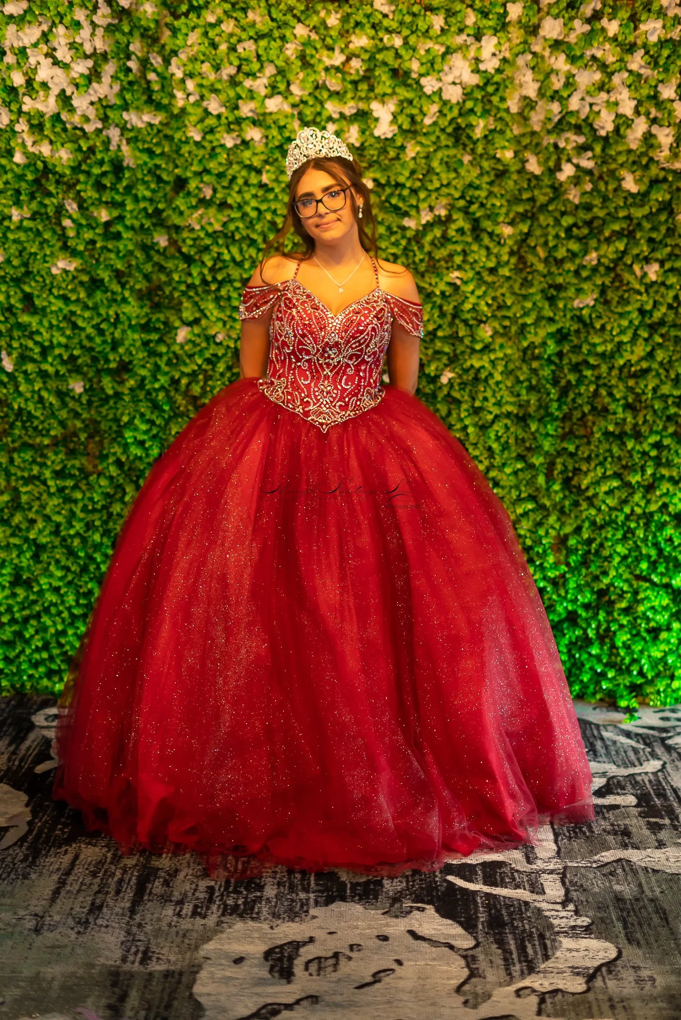 A young woman in a red ball gown with intricate silver embroidery and a full, glittering skirt, wearing a tiara and glasses, posing in front of a green leafy wall.