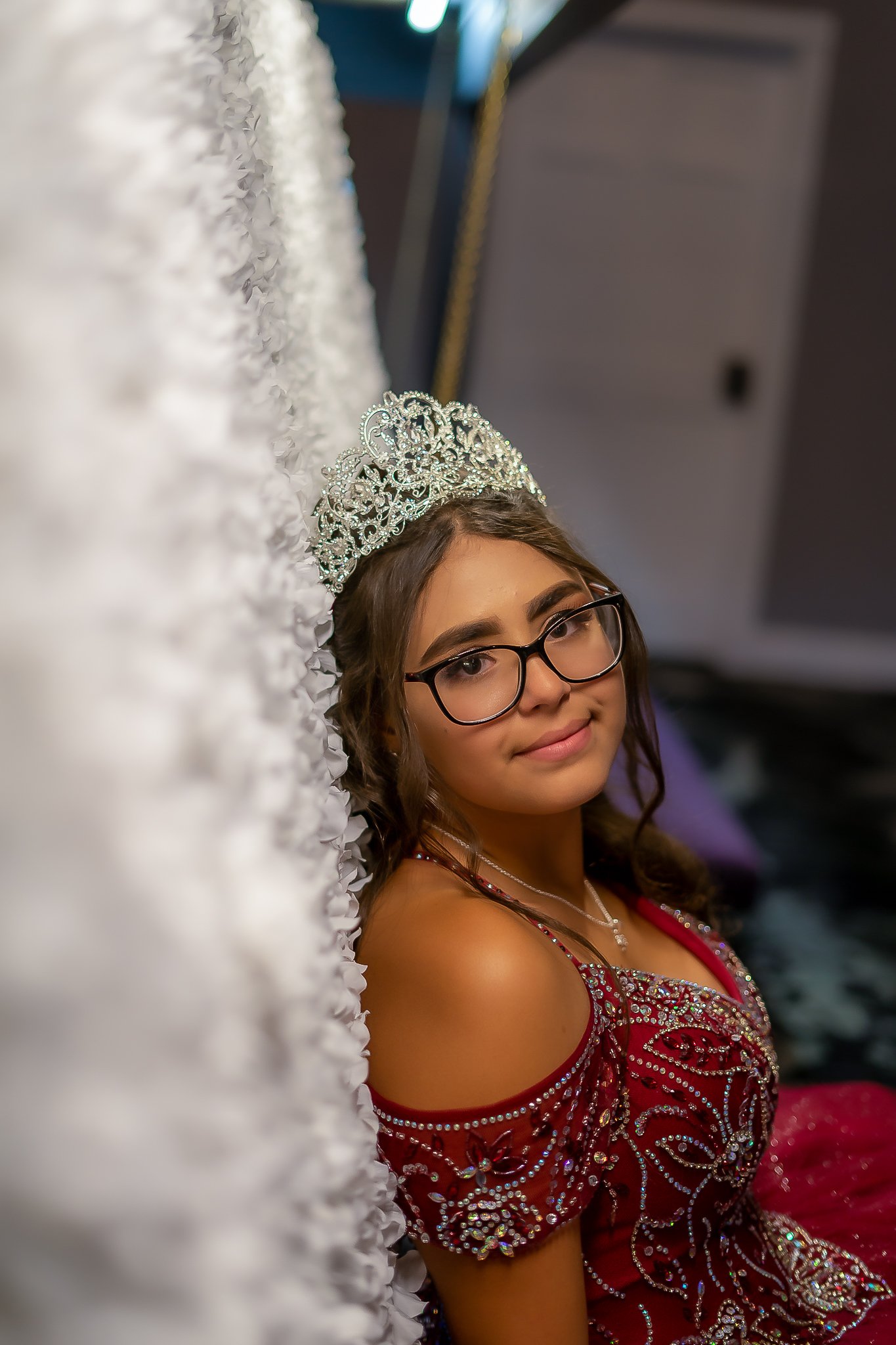 A young girl wearing glasses, a tiara, and a red dress with intricate beadwork and rhinestones, is sitting beside a white textured surface.