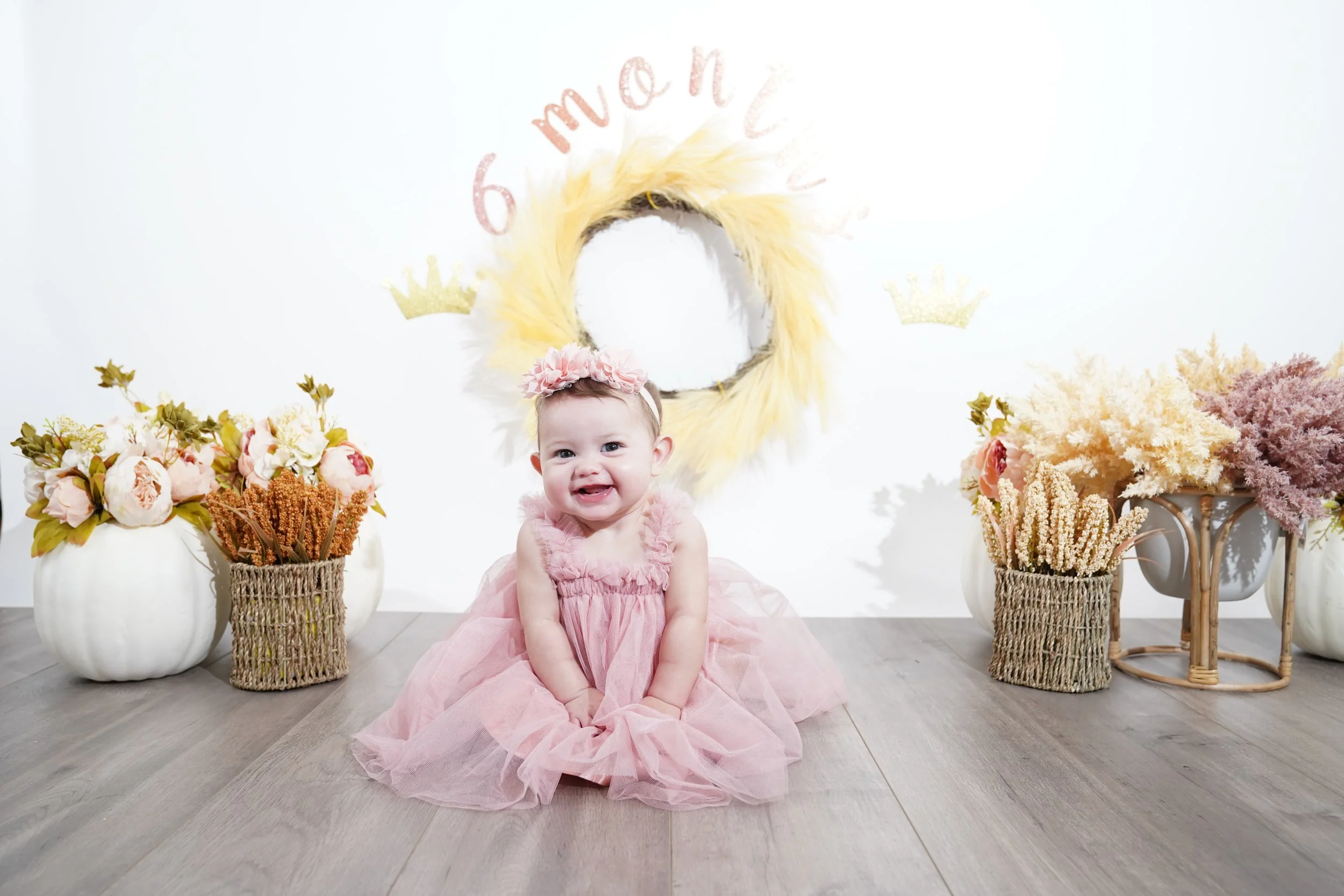 A smiling baby girl sitting on the floor wearing a pink dress and a flower headband, surrounded by floral arrangements in white pots and baskets, with a decorative wall display in the background.