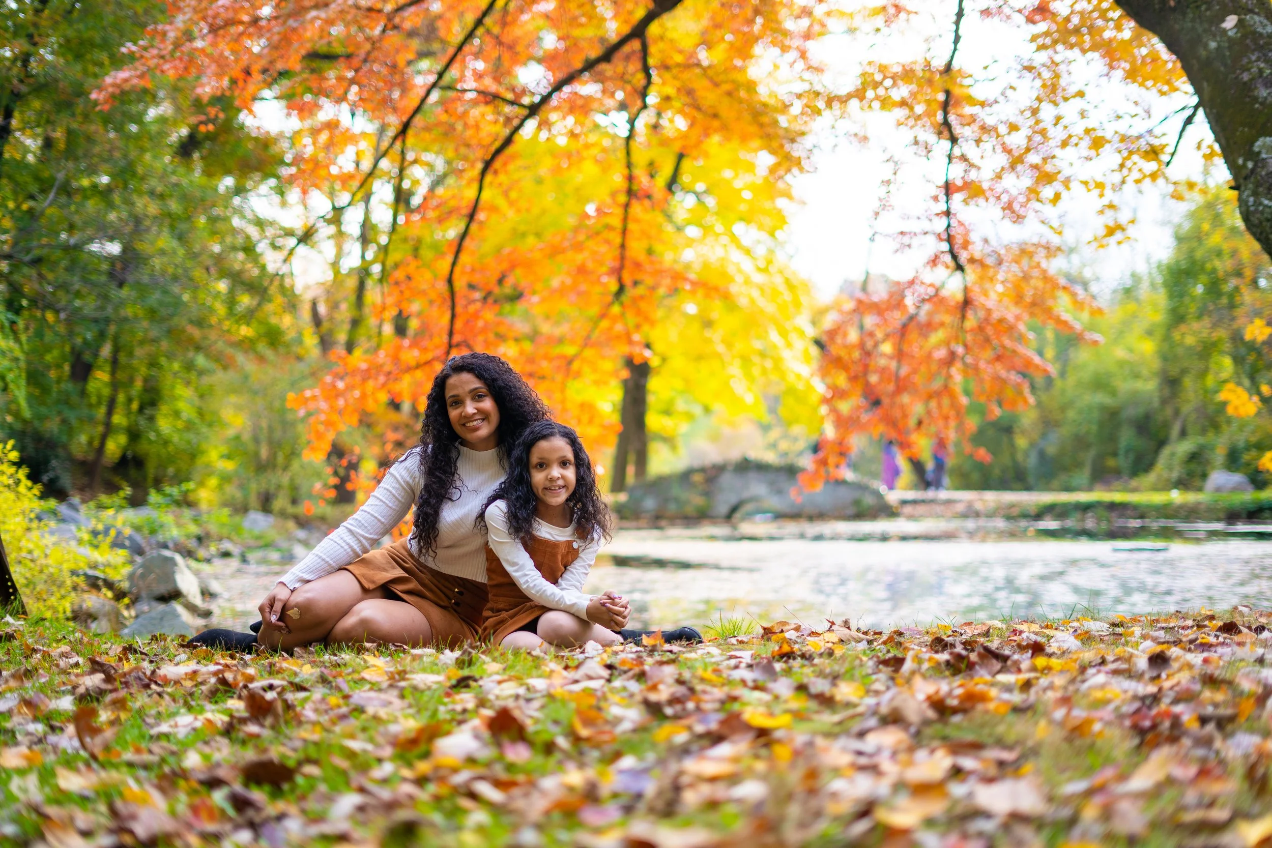A woman and a young girl sitting on the ground in a park surrounded by autumn foliage. The woman has dark curly hair and is smiling, while the girl has long curly hair and is sitting close to her, also smiling. The ground is covered with fallen leave