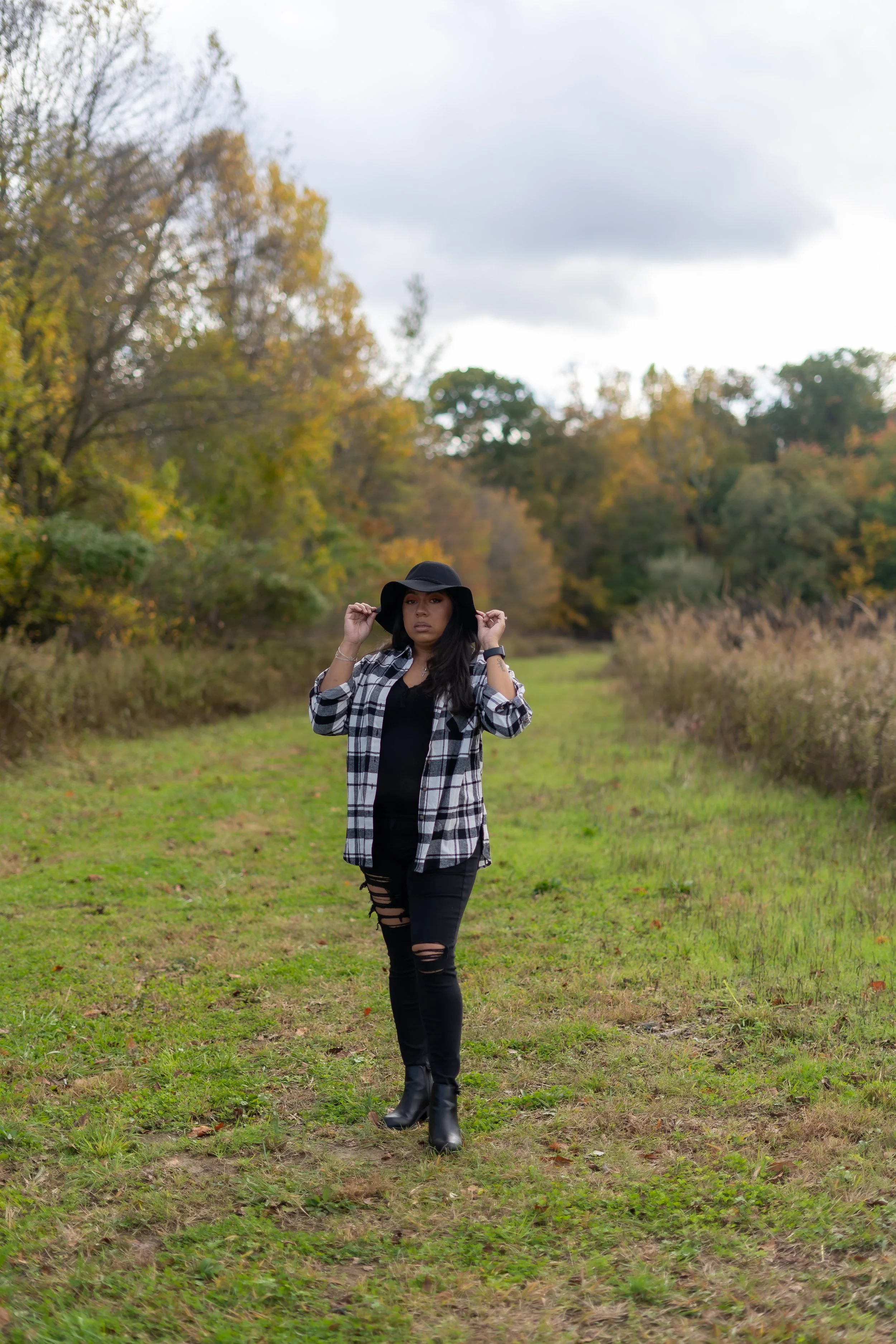A woman standing on a grassy trail in an autumn landscape, holding a black wide-brimmed hat, wearing a black shirt, a plaid shirt, ripped black jeans, and black ankle boots.