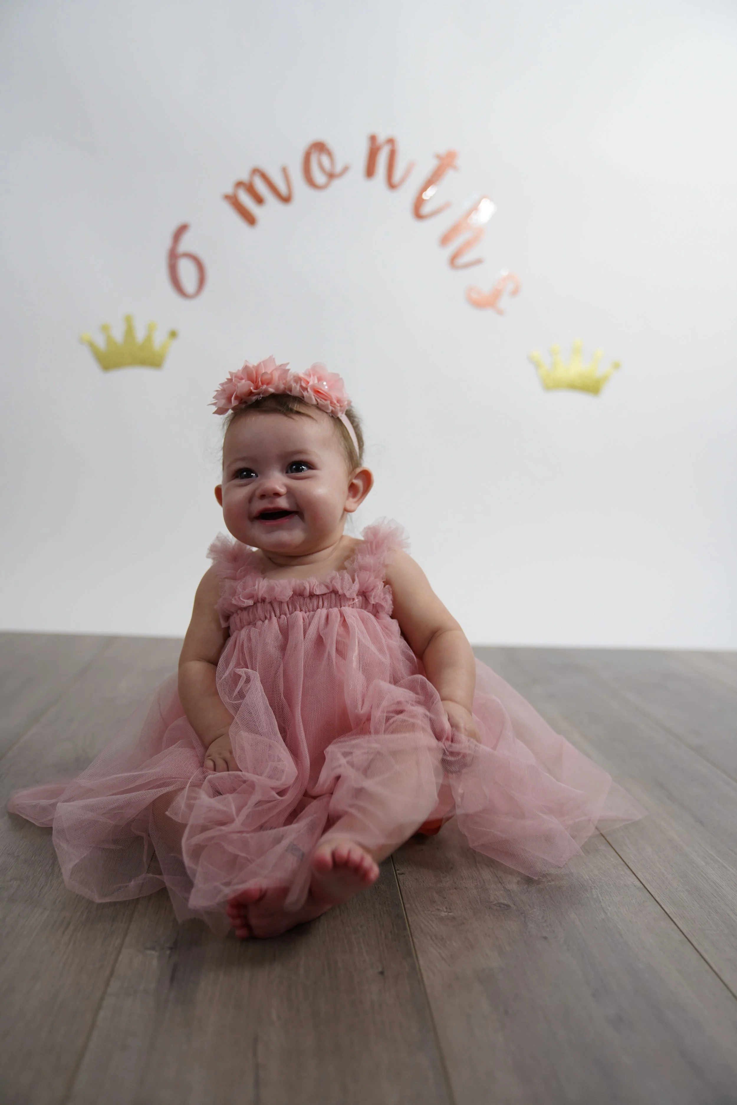 A smiling baby girl wearing a pink tulle dress and a matching pink floral headband, sitting on the floor in front of a white wall decorated with pink lettering that reads 'Moments' and two gold crowns.