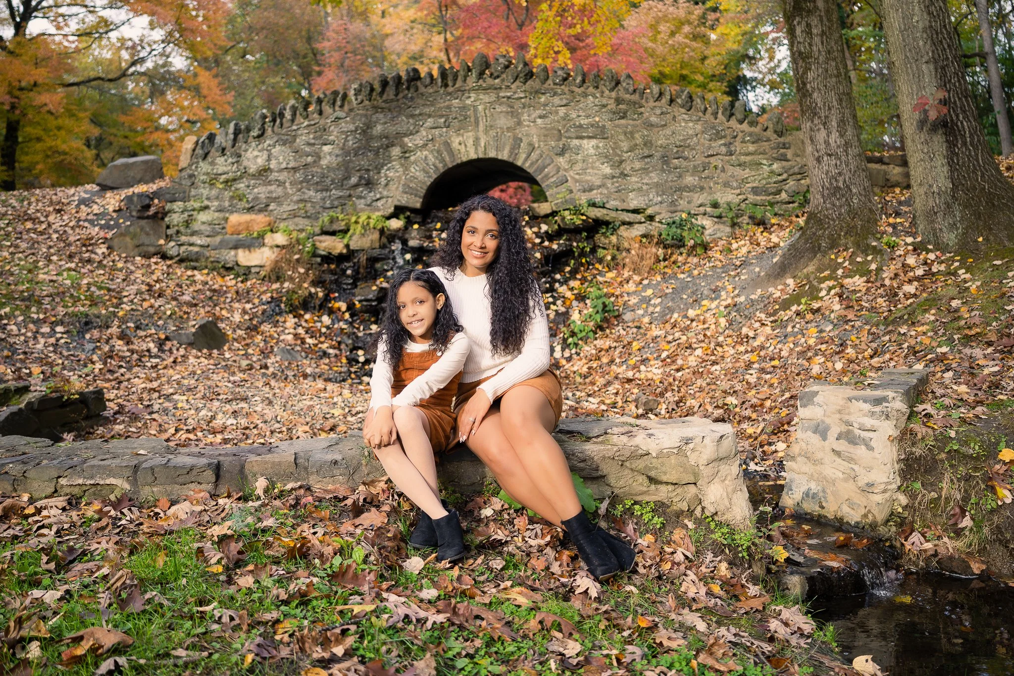 A woman and a young girl sit together on a stone ledge in autumn trees with colorful leaves in the background, near a small creek.