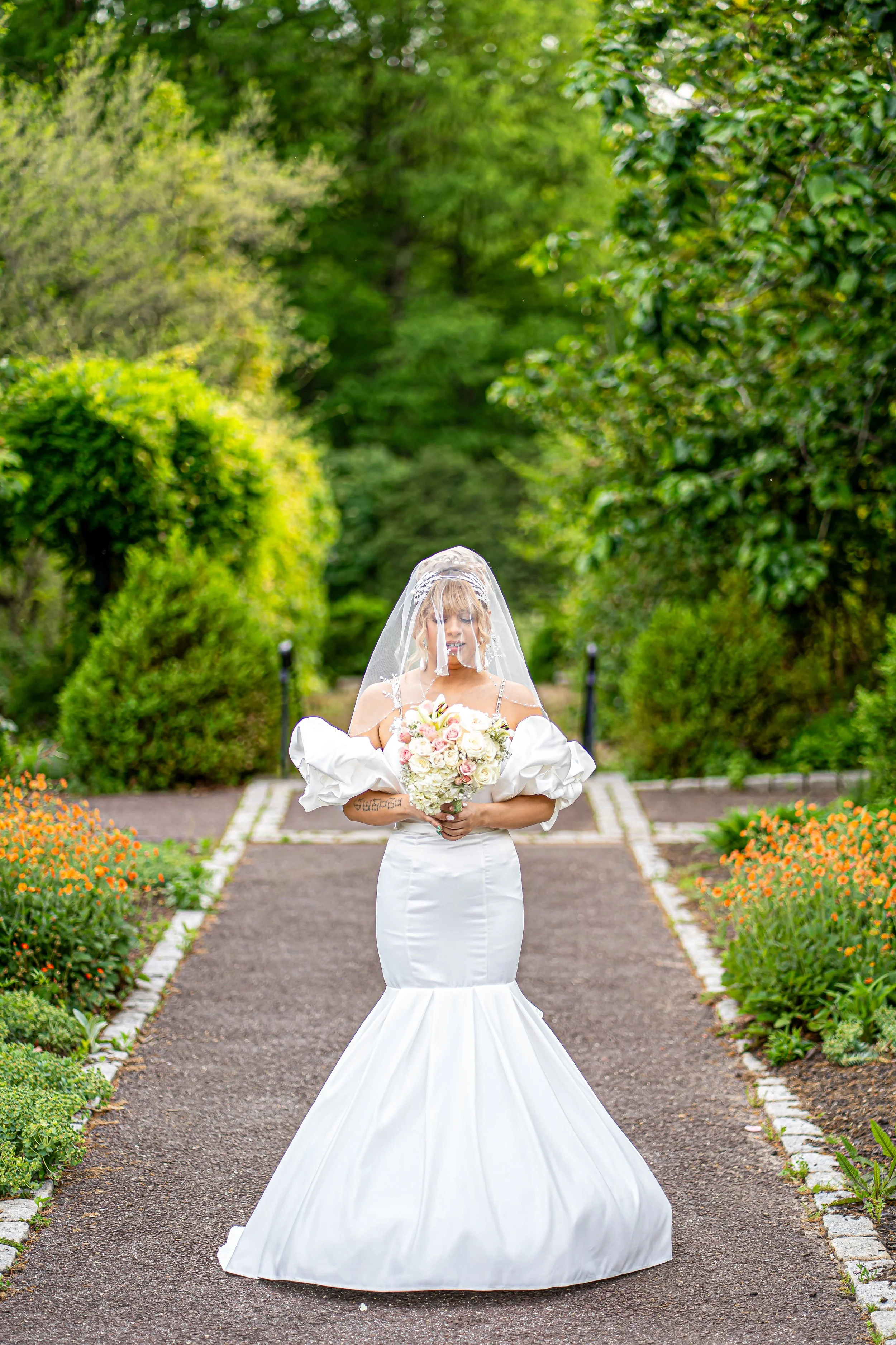 A bride in a white wedding dress and veil standing on a garden path holding a bouquet of white and pink roses, surrounded by greenery and flower beds.
