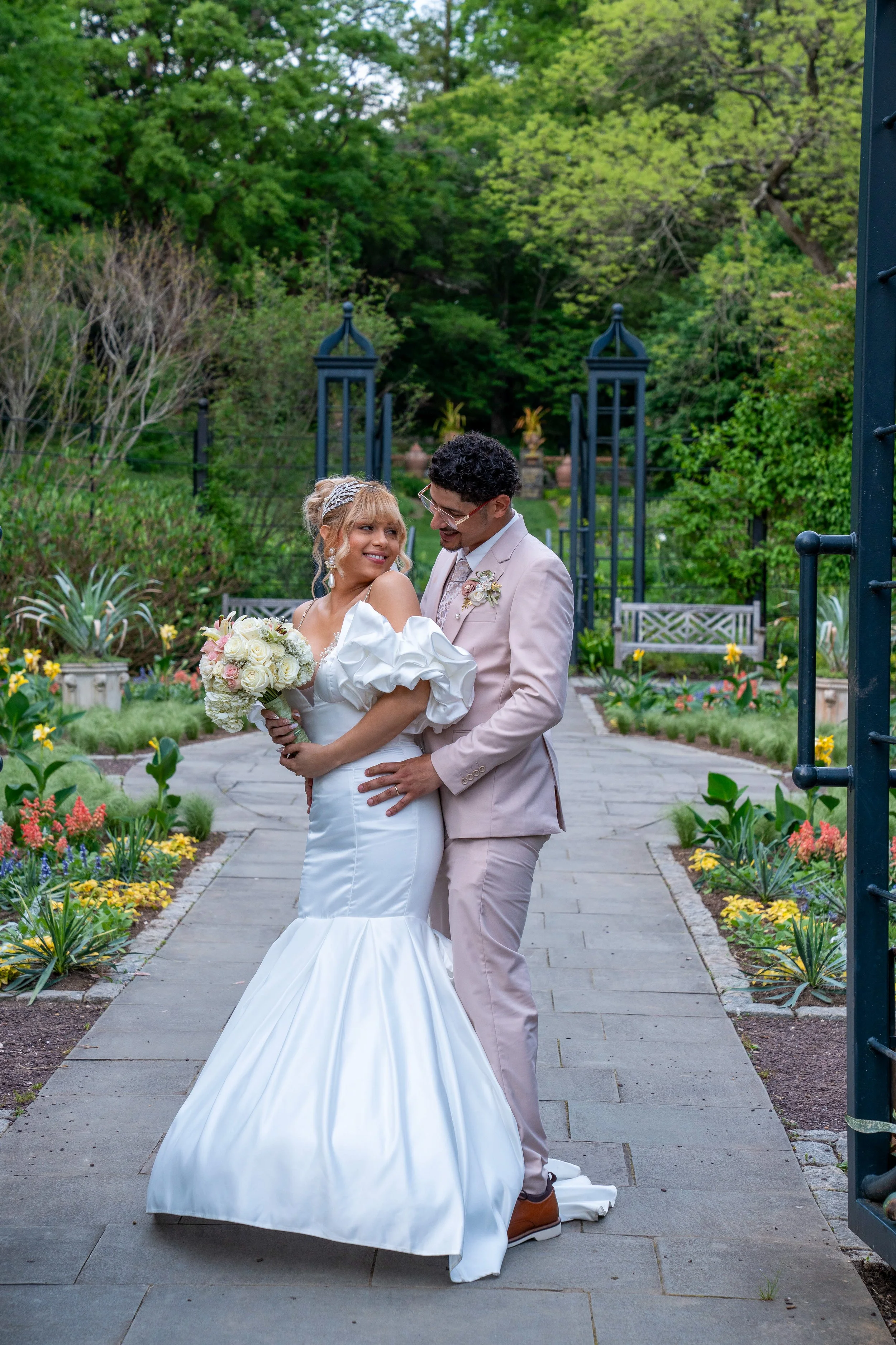 A bride and groom smiling and looking at each other in a garden on their wedding day, surrounded by greenery and colorful flowers, with a paved path and garden benches visible.