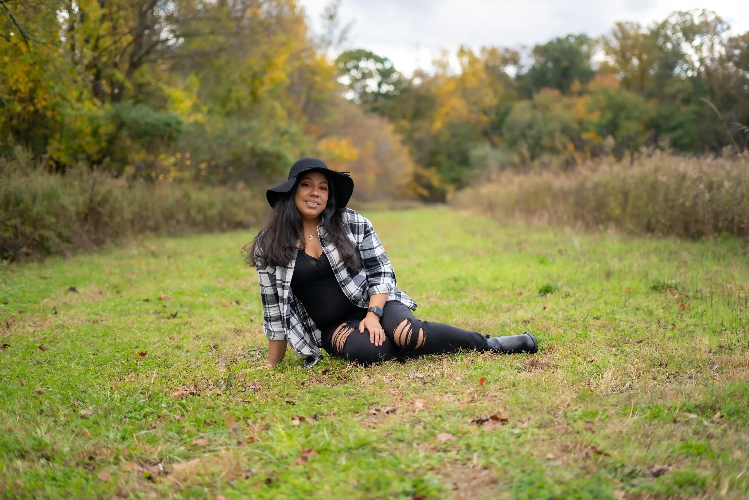 A young woman with long dark hair sitting on grass in a park during autumn, wearing a black hat, plaid shirt, ripped black jeans, and black boots, smiling at the camera with trees displaying fall foliage in the background.