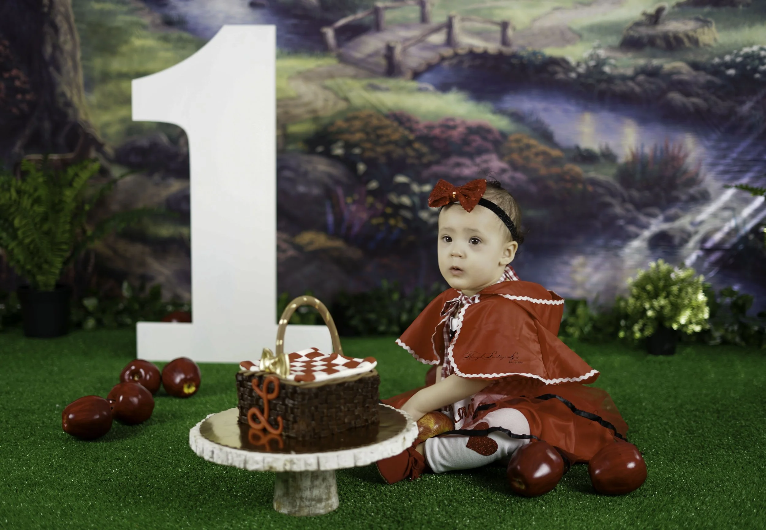 A young girl dressed in a red cape with a red bow headband sitting on green artificial grass near a chocolate birthday cake with red chocolate numbers, red apples, and a nature-themed backdrop.