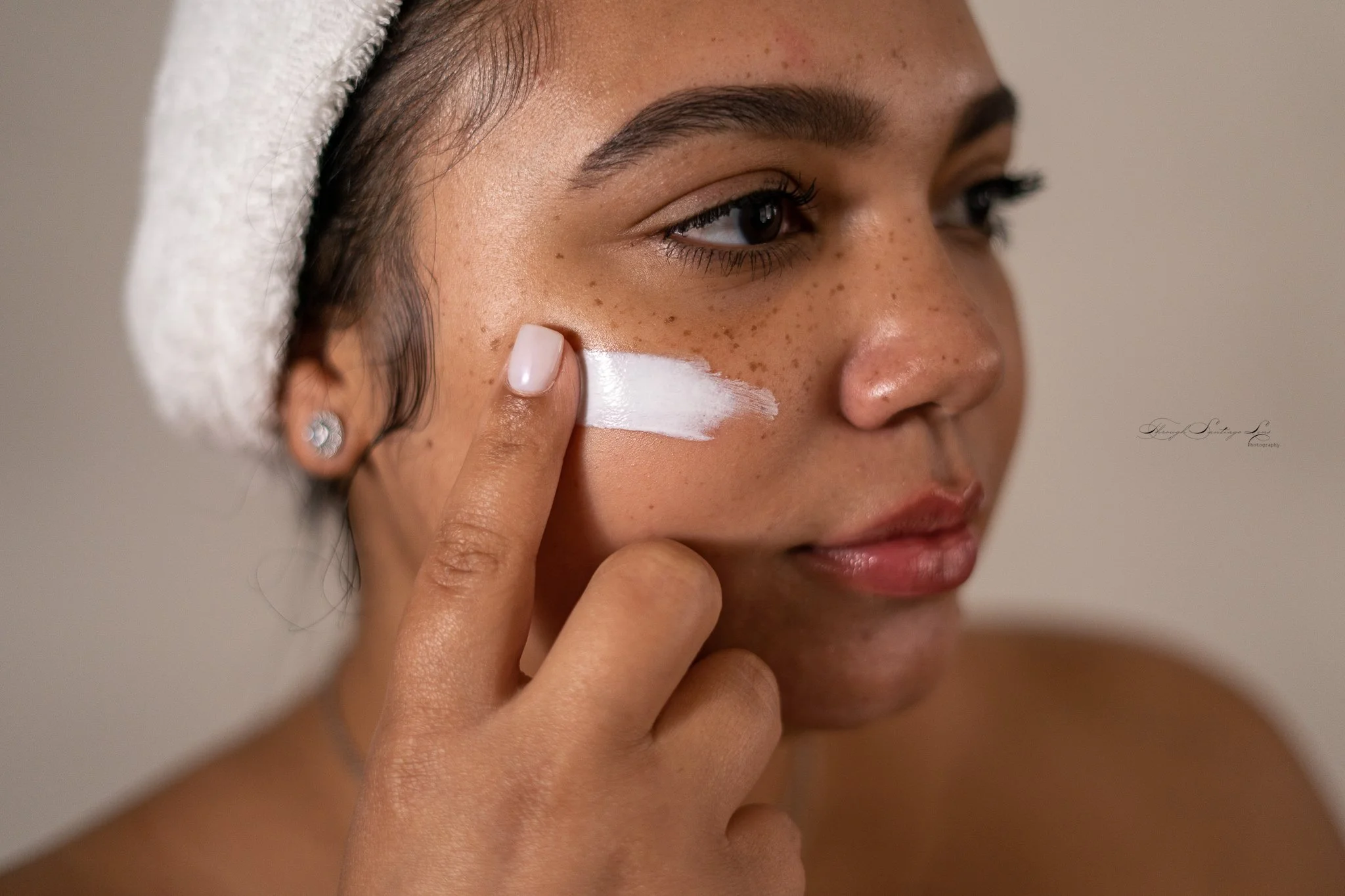 A woman with brown skin and freckles applying sunscreen to her face, with a towel wrapped around her head.