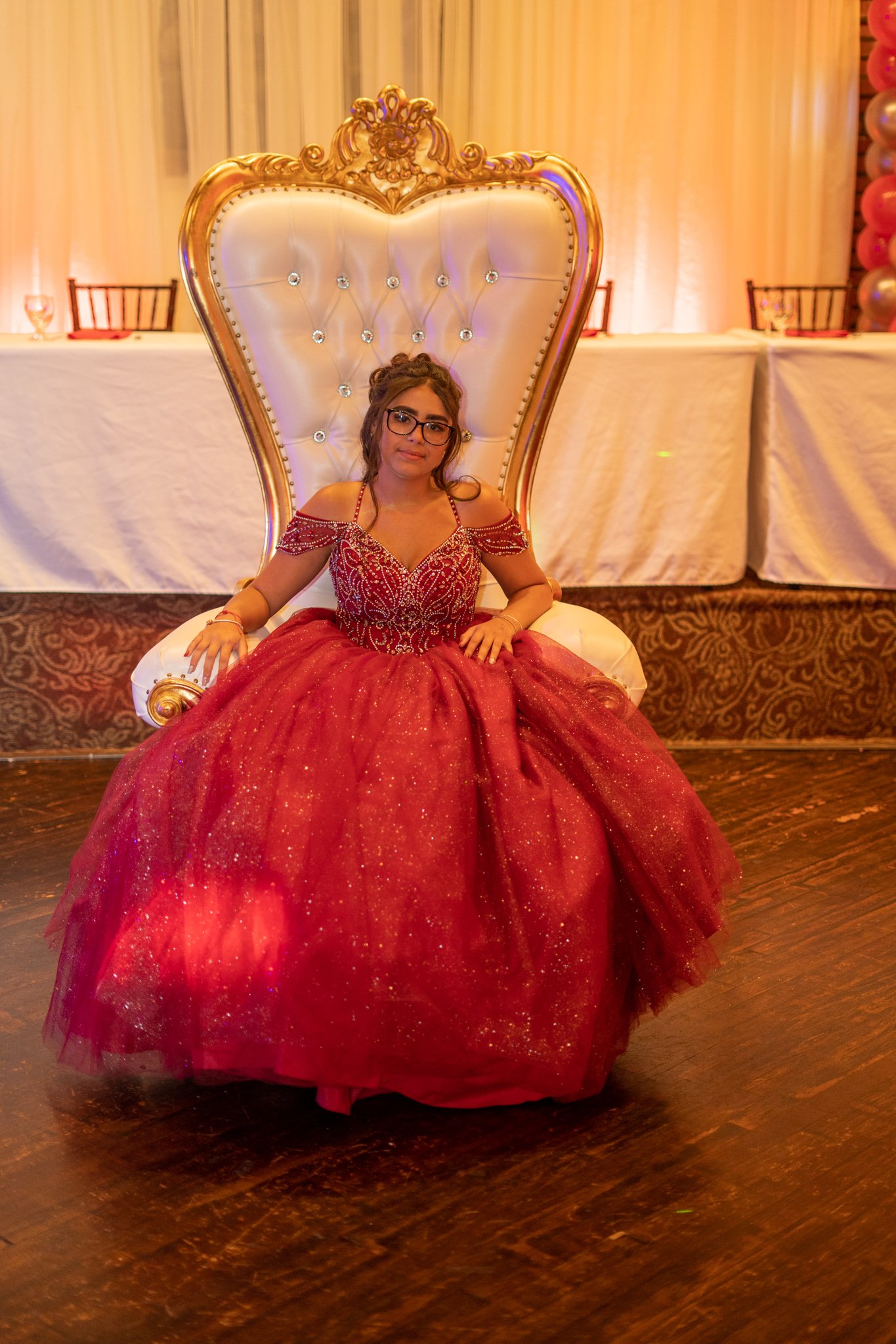 Young woman sitting on a decorated throne at a celebration, wearing a red, glittery gown with off-shoulder design and glasses, with a detailed backdrop and balloons on the side.