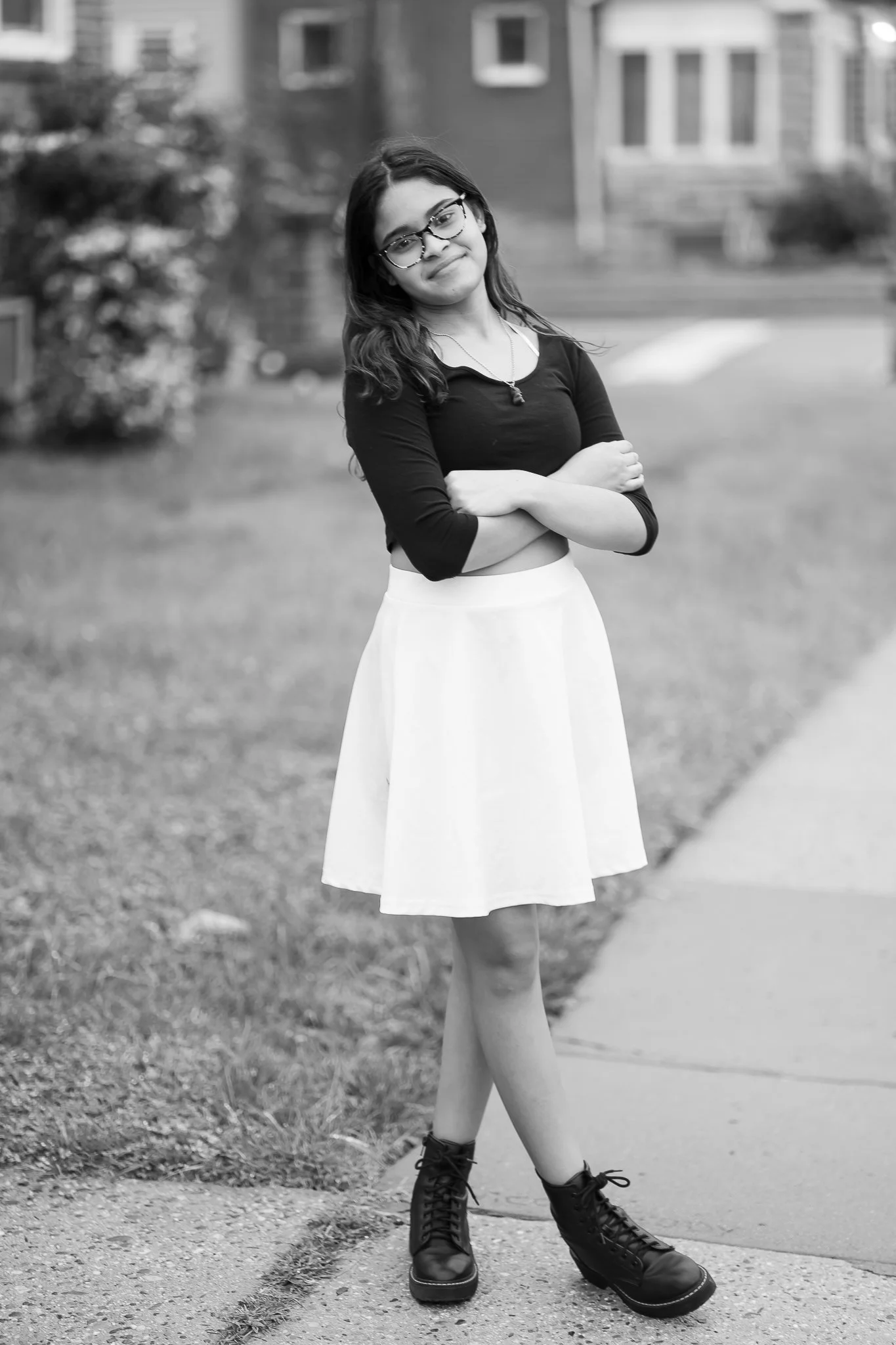 A young woman standing outdoors on a sidewalk wearing a black top, a white skirt, and combat boots, with her arms crossed and a slight smile. She has long wavy hair, glasses, and is wearing a necklace, with a blurred residential background.