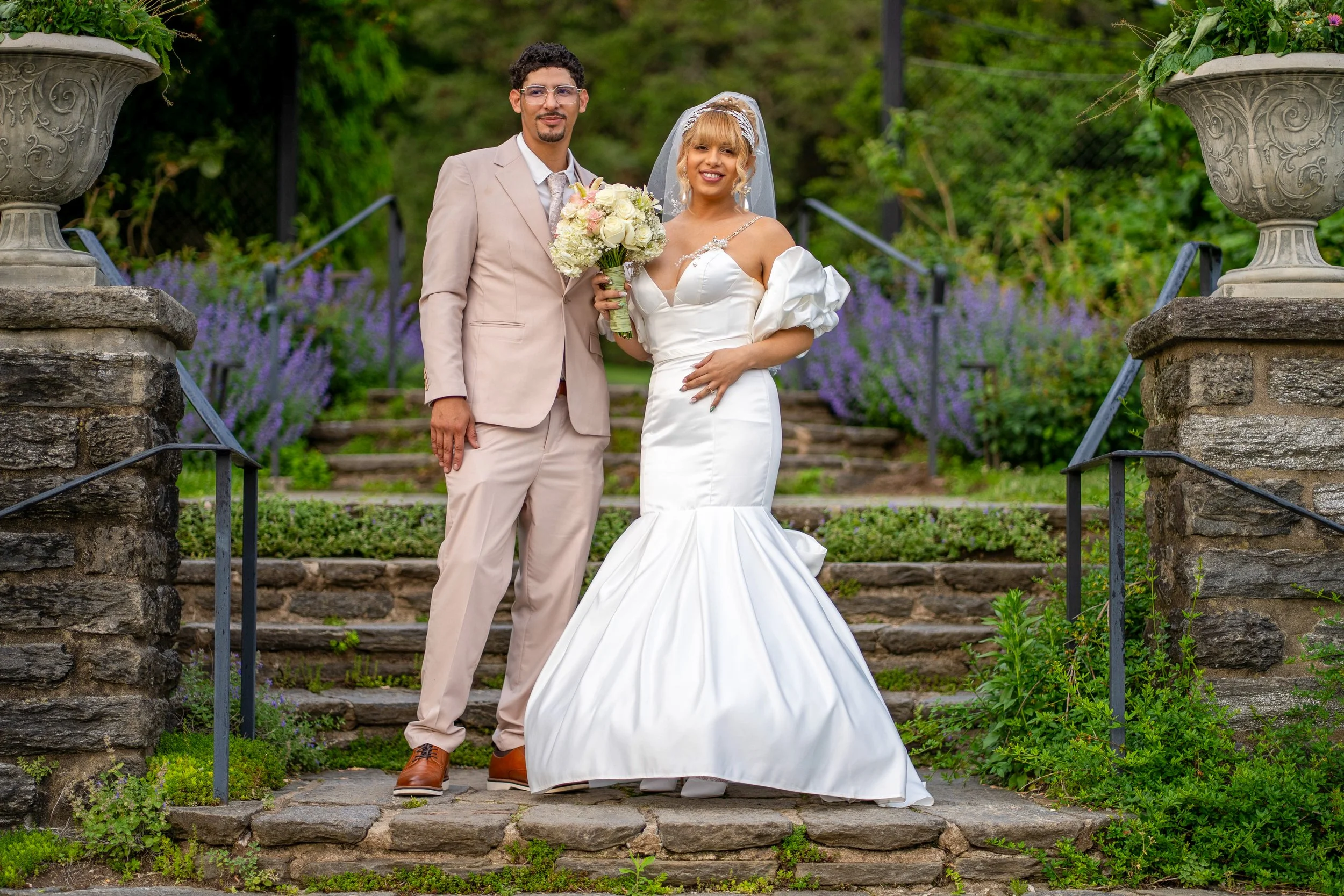 A bride and groom standing on stone steps in a garden, surrounded by green trees and purple flowers. The bride is holding a bouquet of white and pink roses and wearing a strapless white wedding dress with puffed sleeves and a veil. The groom is dress