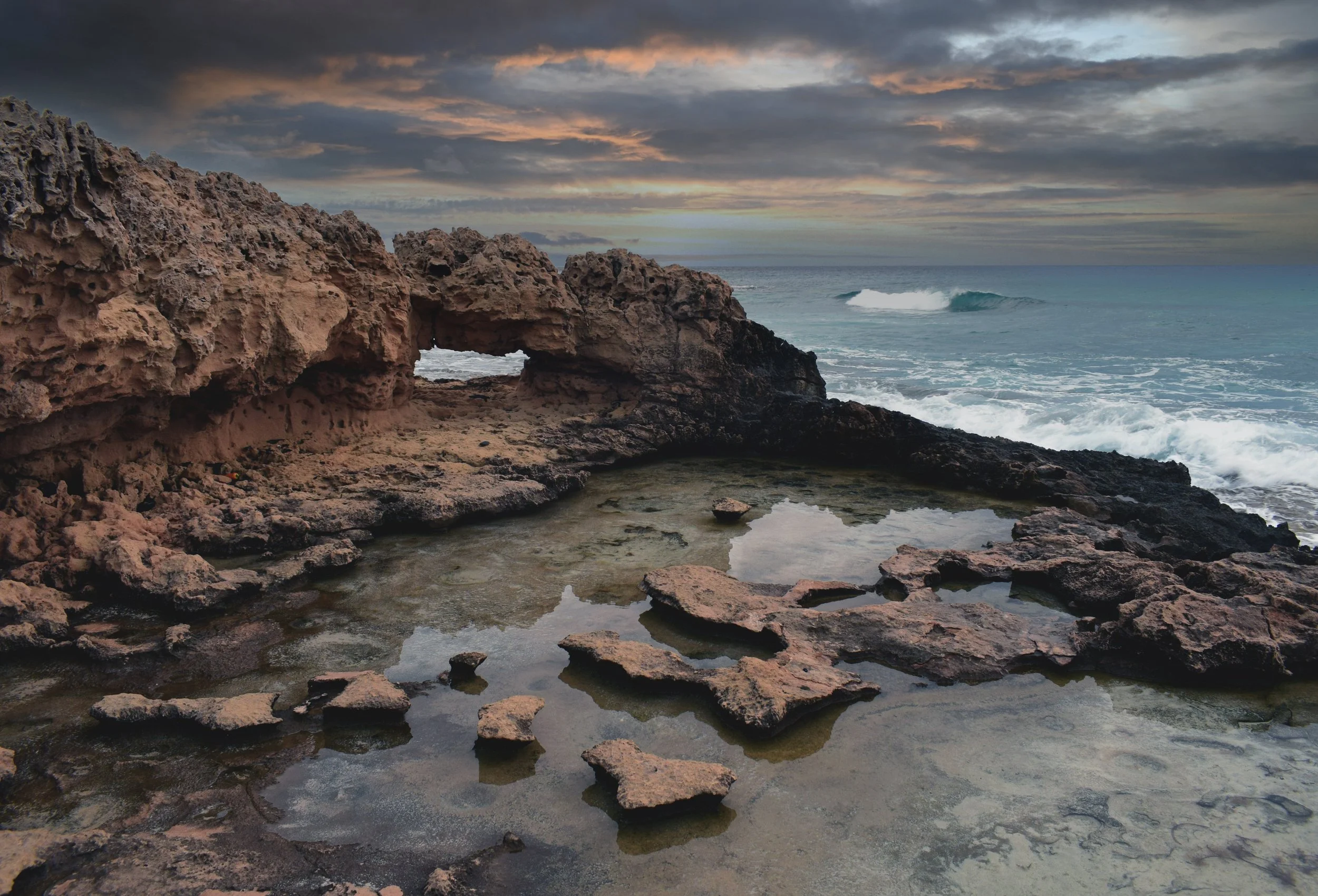 Sunset over rocky shoreline with waves crashing near the coast, dark clouds in the sky.