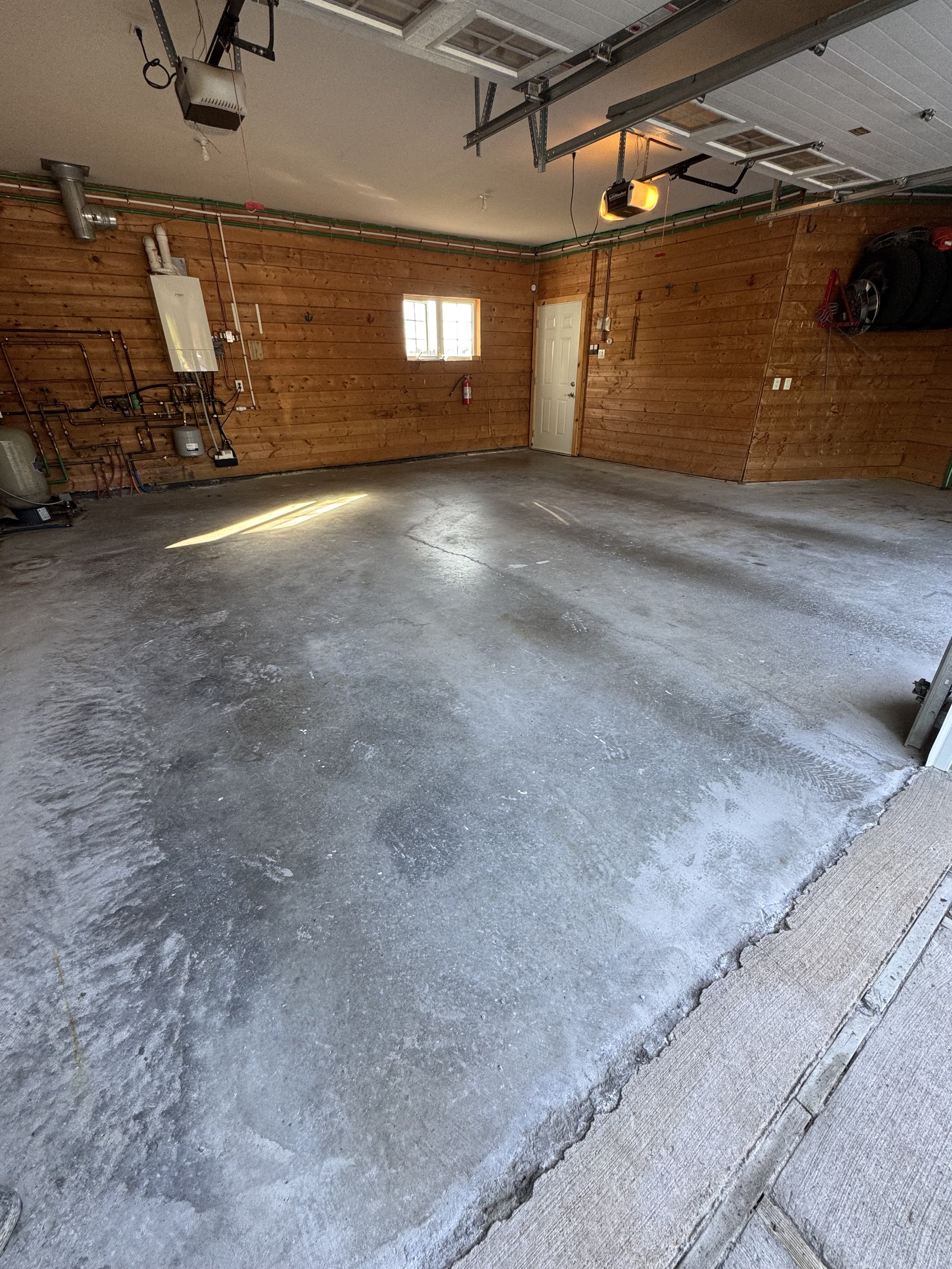 Empty garage with concrete floor, wood-paneled walls, a window, and a door. Garage door openers and some tools are visible, along with a tire rack on the wall.