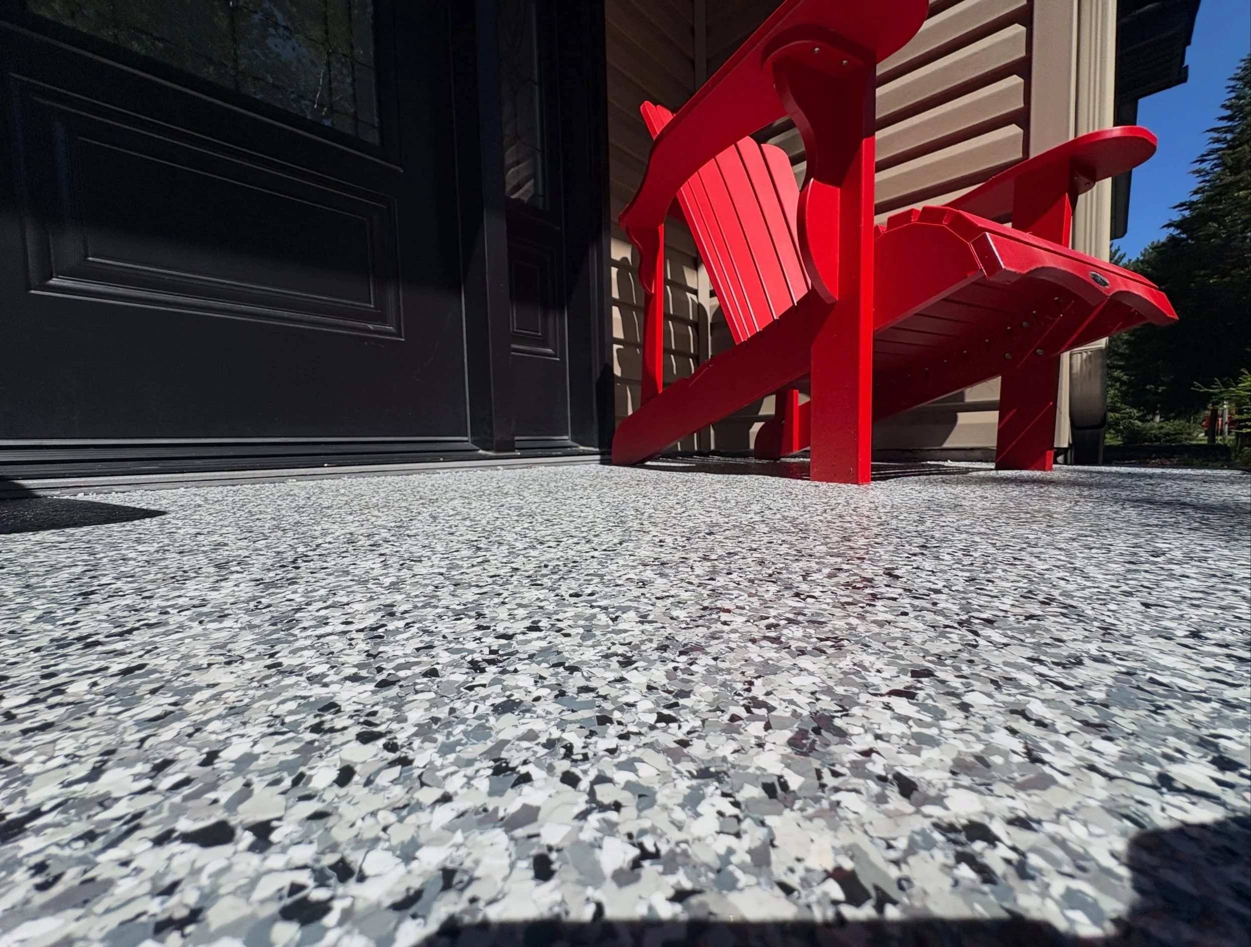 A red Adirondack chair on a porch resting on terrazzo flooring, with a black door and window, beige siding, and trees in the background.