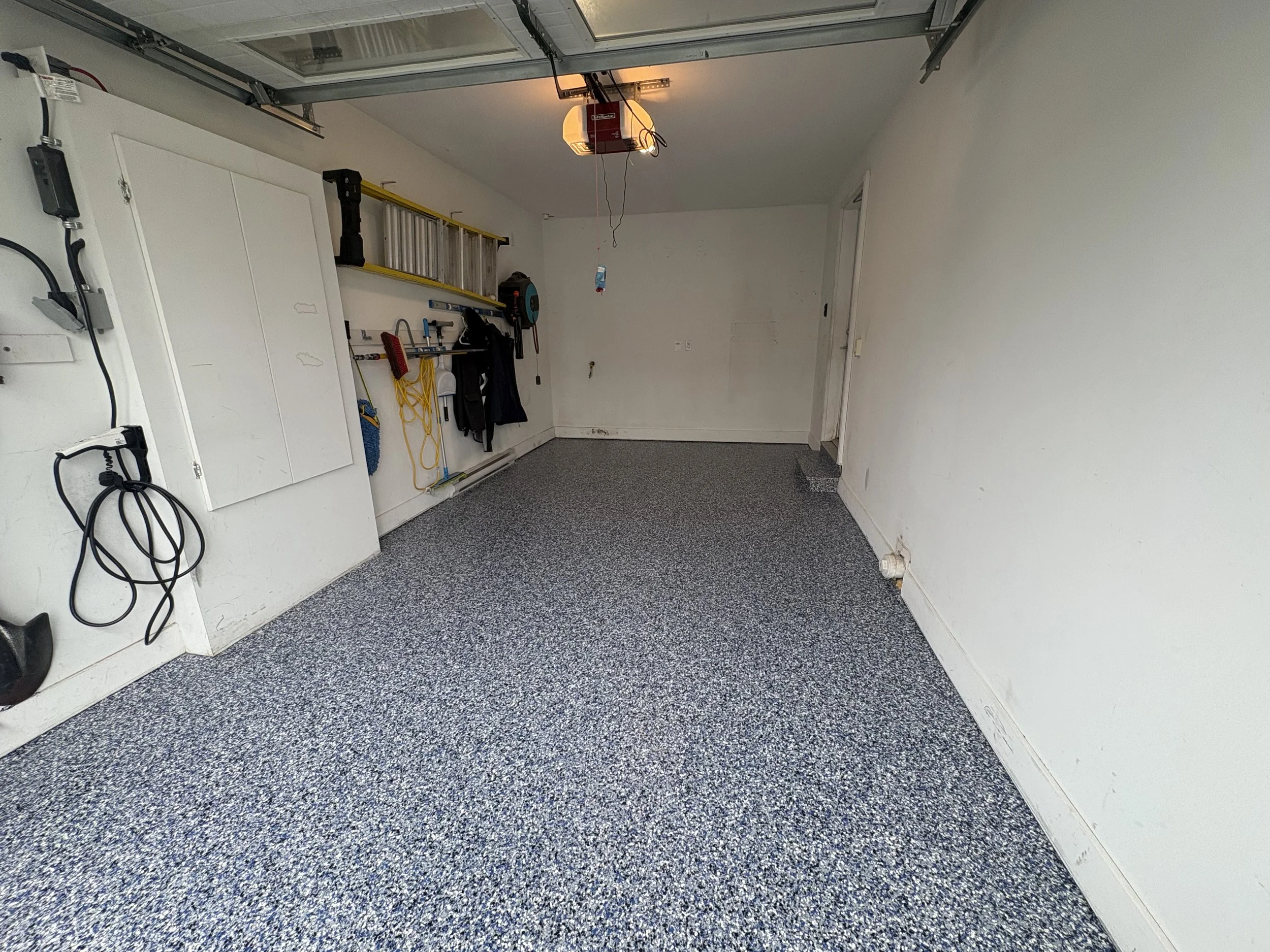 An empty garage with gray speckled floor, white walls, and a partially open garage door. There are items hanging on the wall including a ladder, a broom, and some tools, with some electrical cords and chargers visible.