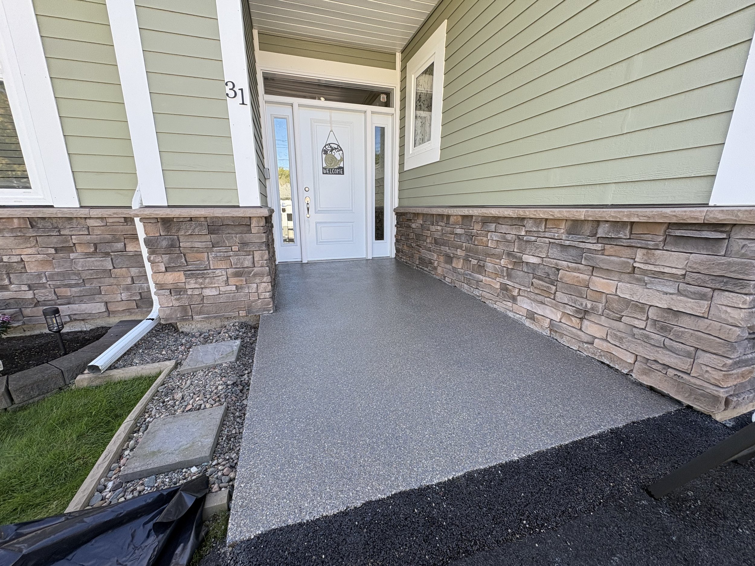 A house entrance with a white door, flanked by two side windows, and the house number 31. The house has green siding with white trim and a stone foundation. The porch area features a gray concrete floor and a landscaped border with grass, stones, and