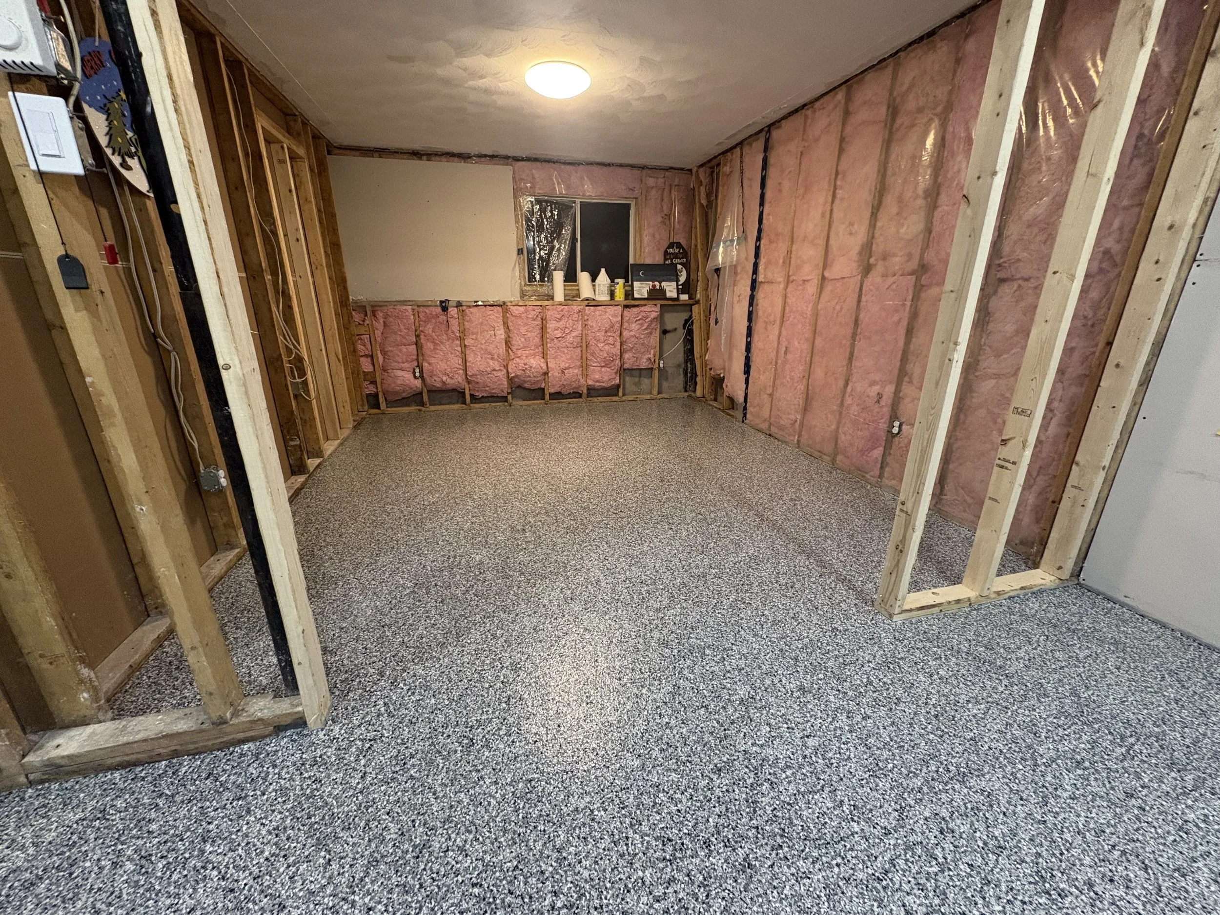 An unfinished room with exposed wooden studs and pink insulation. The ceiling has a light fixture, and there's a small window on the back wall. The floor is covered with speckled gray carpet.