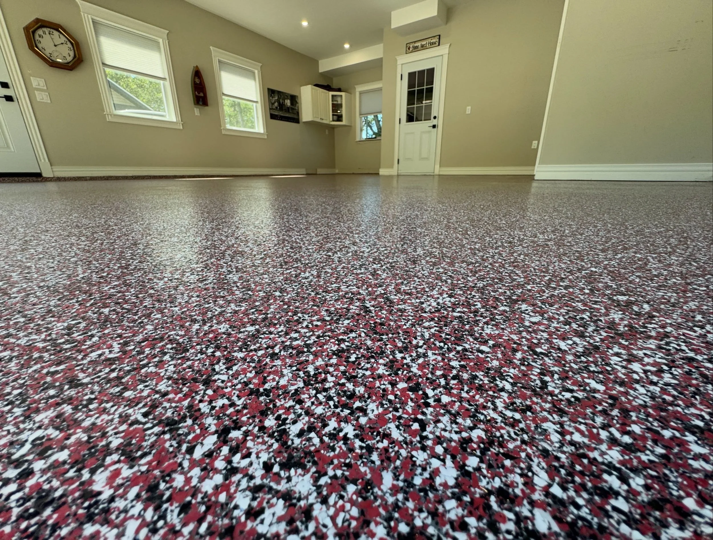 Empty living room with beige walls, three windows with blinds, a clock on the wall, and a door leading outside; epoxy floor with red, black, and white speckled pattern.