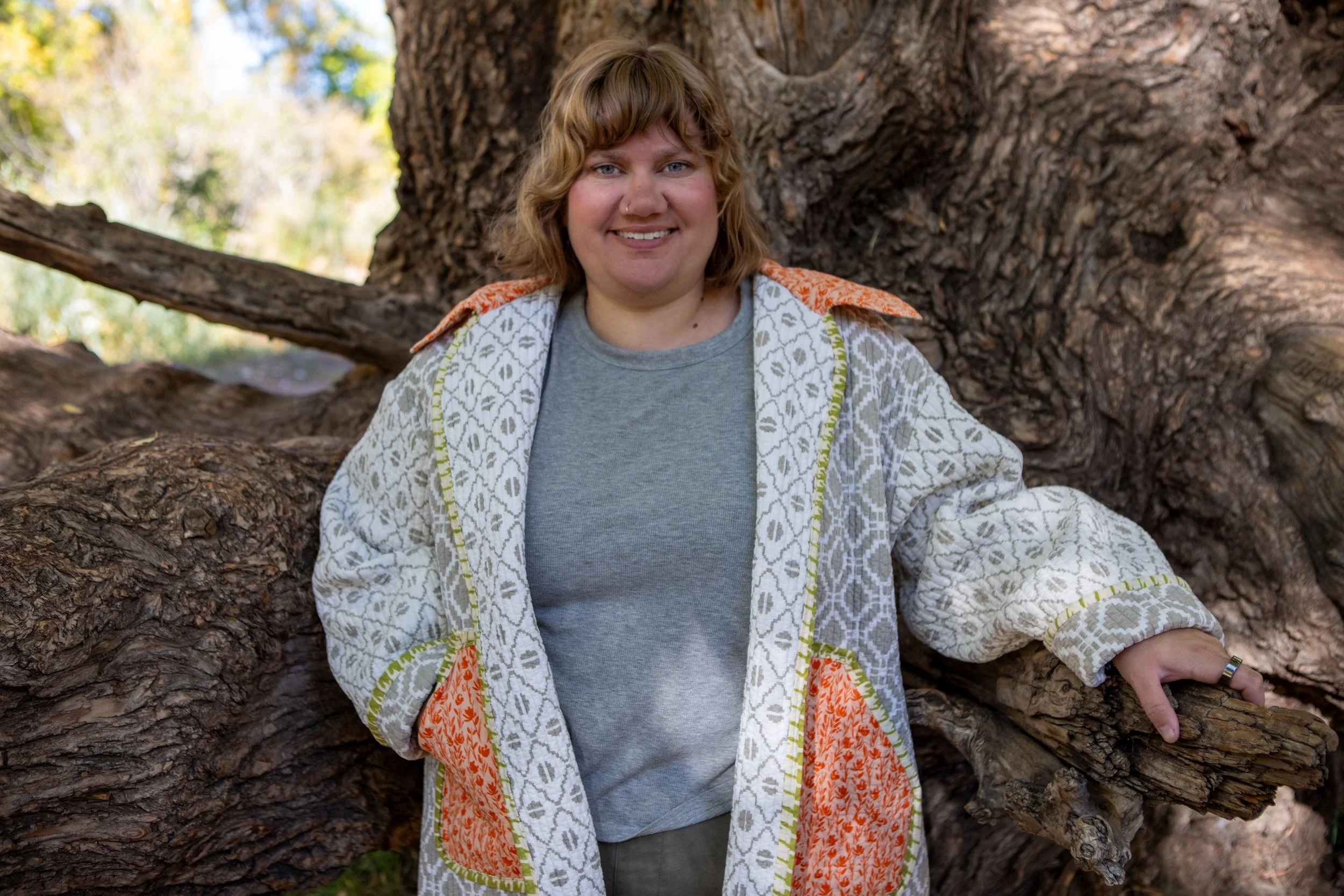 Courtney Allen with short wavy light brown hair smiling outdoors, sitting next to a large tree.