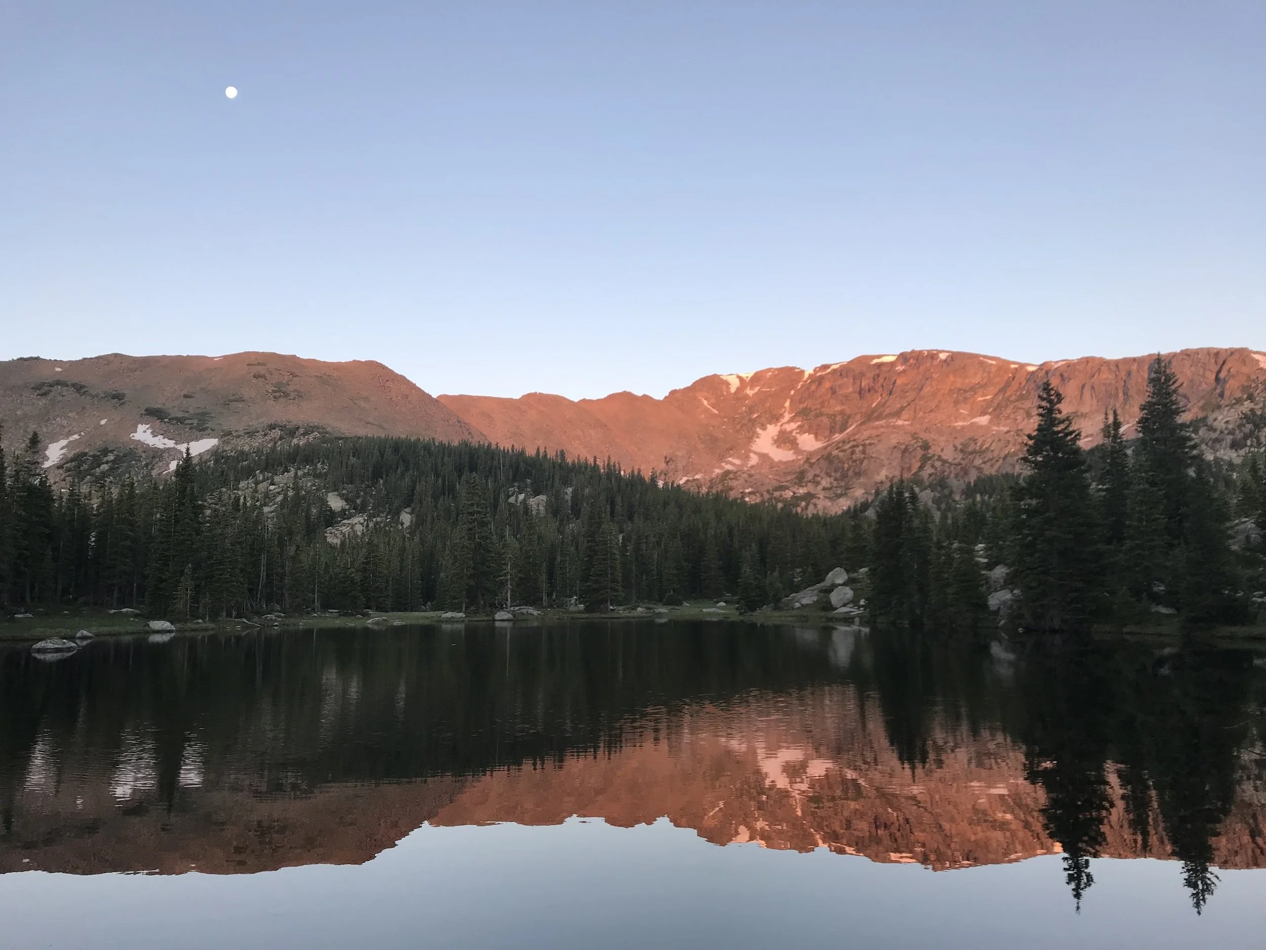 Scenic view of mountain range in Colorado reflecting in a lake at sunset, with trees in the foreground and the moon visible in the sky.