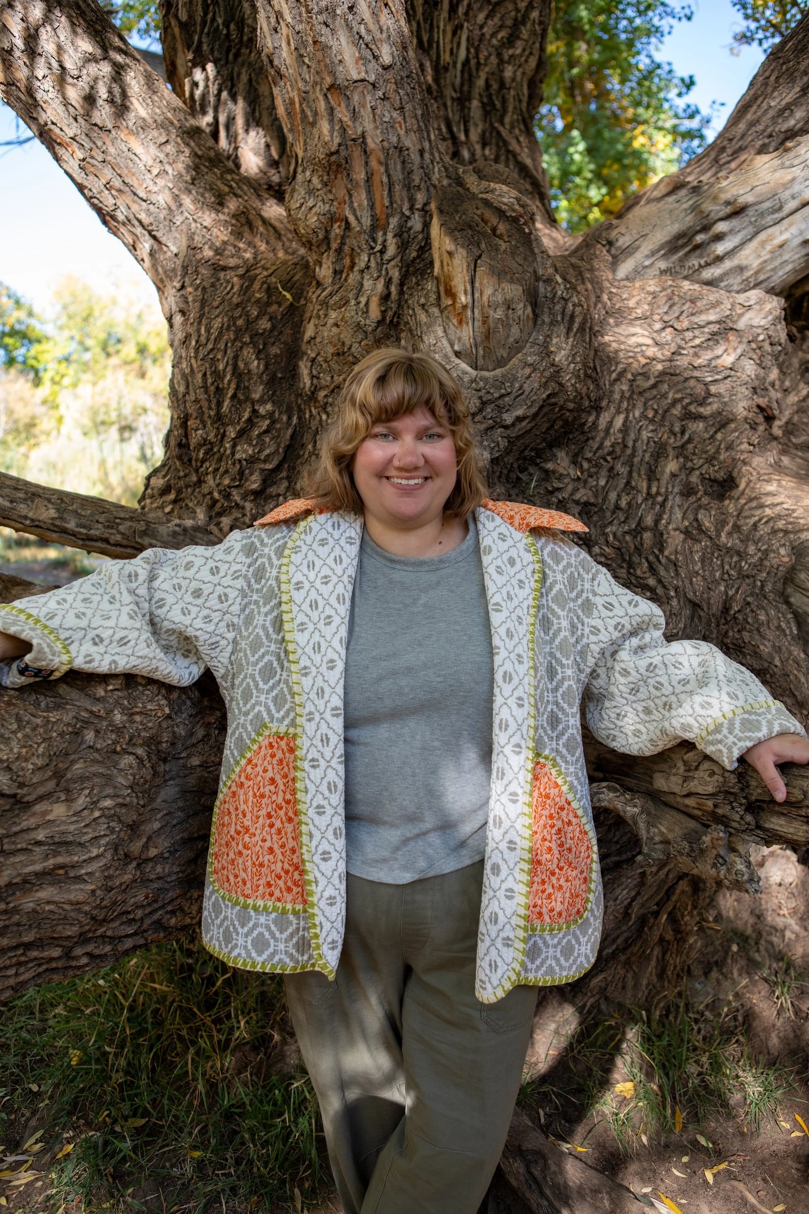 Courtney Allen with curly short hair smiling and posing with their arms spread out, standing in front of a large, thick tree trunk outdoors with green leaves and a partly cloudy sky in the background.