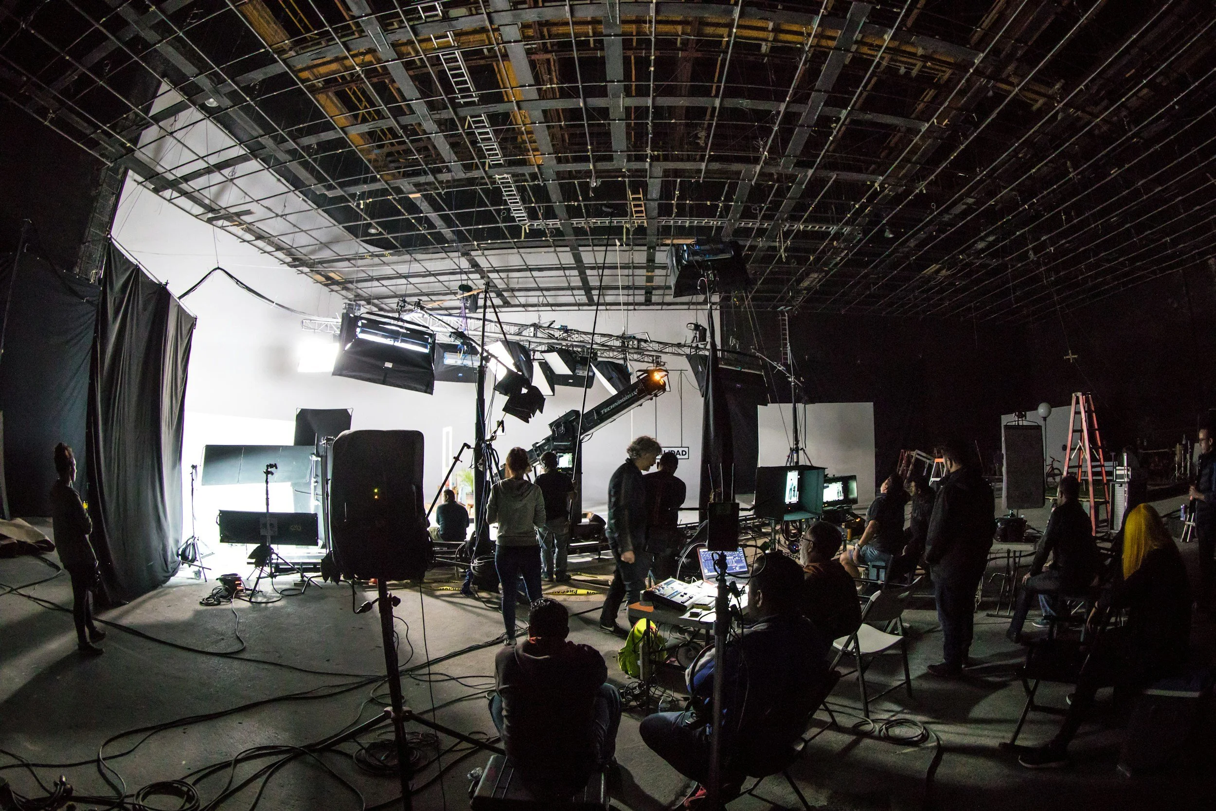 Indoor studio with crew members preparing for filming or photography, with equipment, lighting, and a white backdrop.