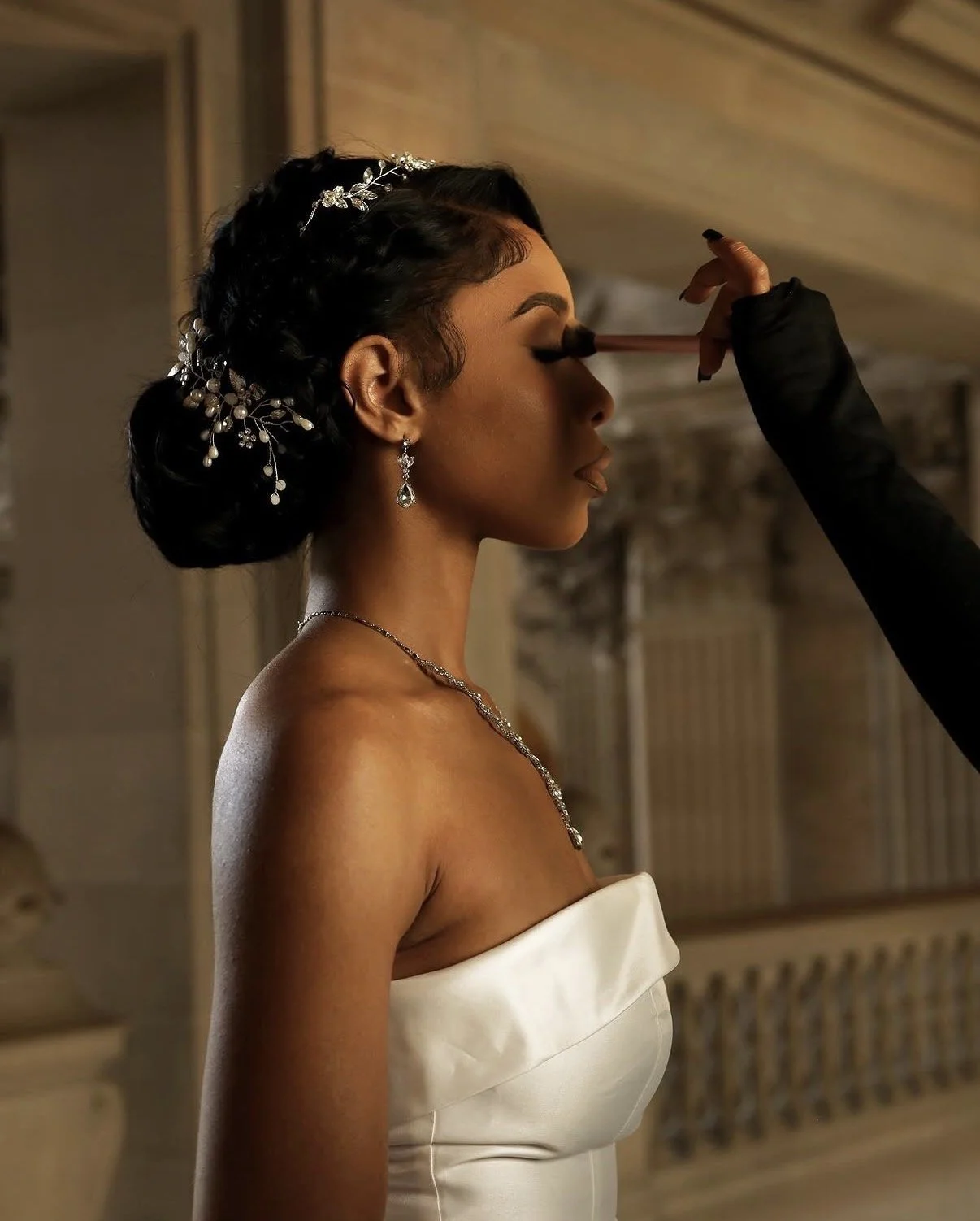Bridesmaid getting makeup applied in a wedding dress with jewelry and an elegant updo hairstyle.