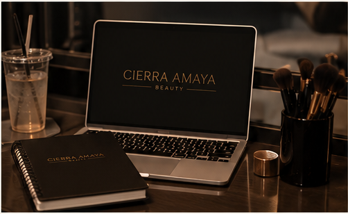 A laptop with a screen displaying 'CIERRA AMAYA BEAUTY', a spiral notebook with the same text, a container with makeup brushes, and a drink with a straw on a wooden table.