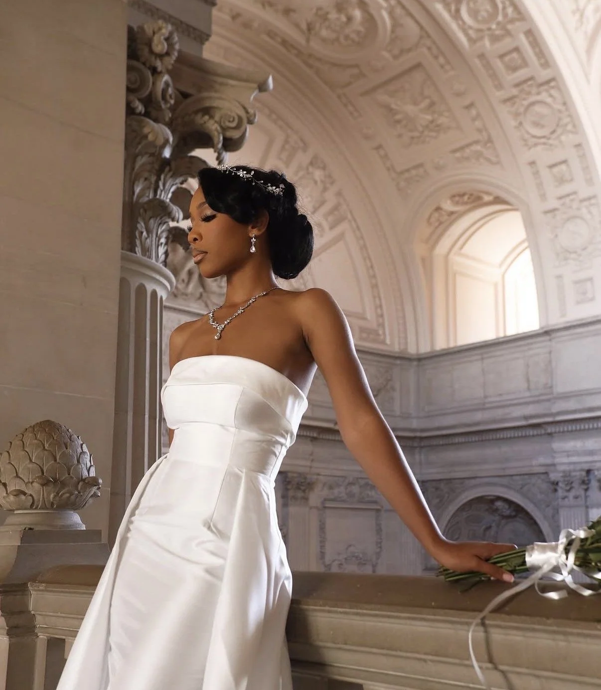A woman in a white wedding gown with jewelry, standing in an ornate, classical-style hall with high arched ceilings and intricate architectural details.