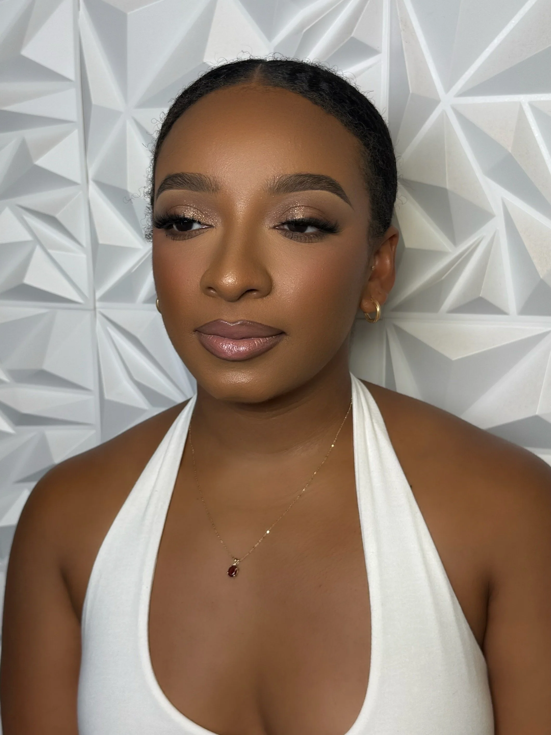 A woman with dark skin and short black hair styled close to her head, wearing eye makeup with gold and black eyeshadow, gold earrings, a delicate gold necklace with a pendant, and a white sleeveless top, standing against a textured white background.