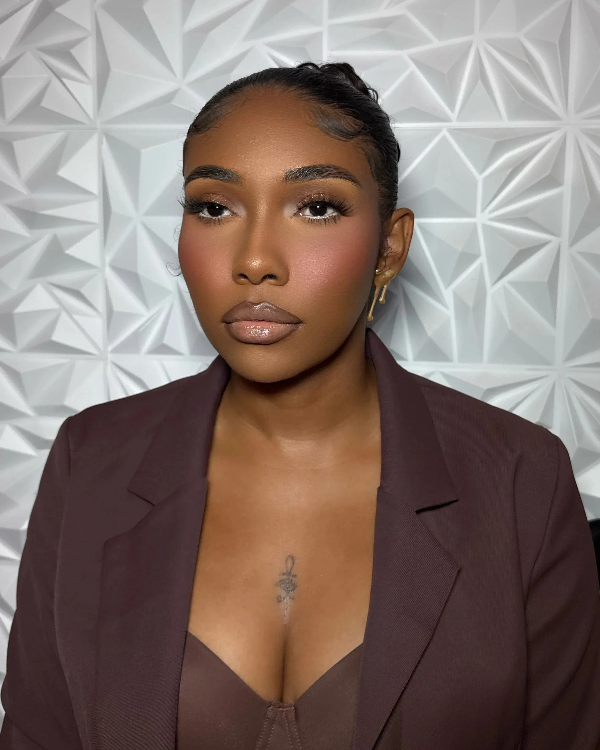 A woman with dark skin and makeup, wearing gold earrings and a dark blazer, poses against a white geometric wall background.