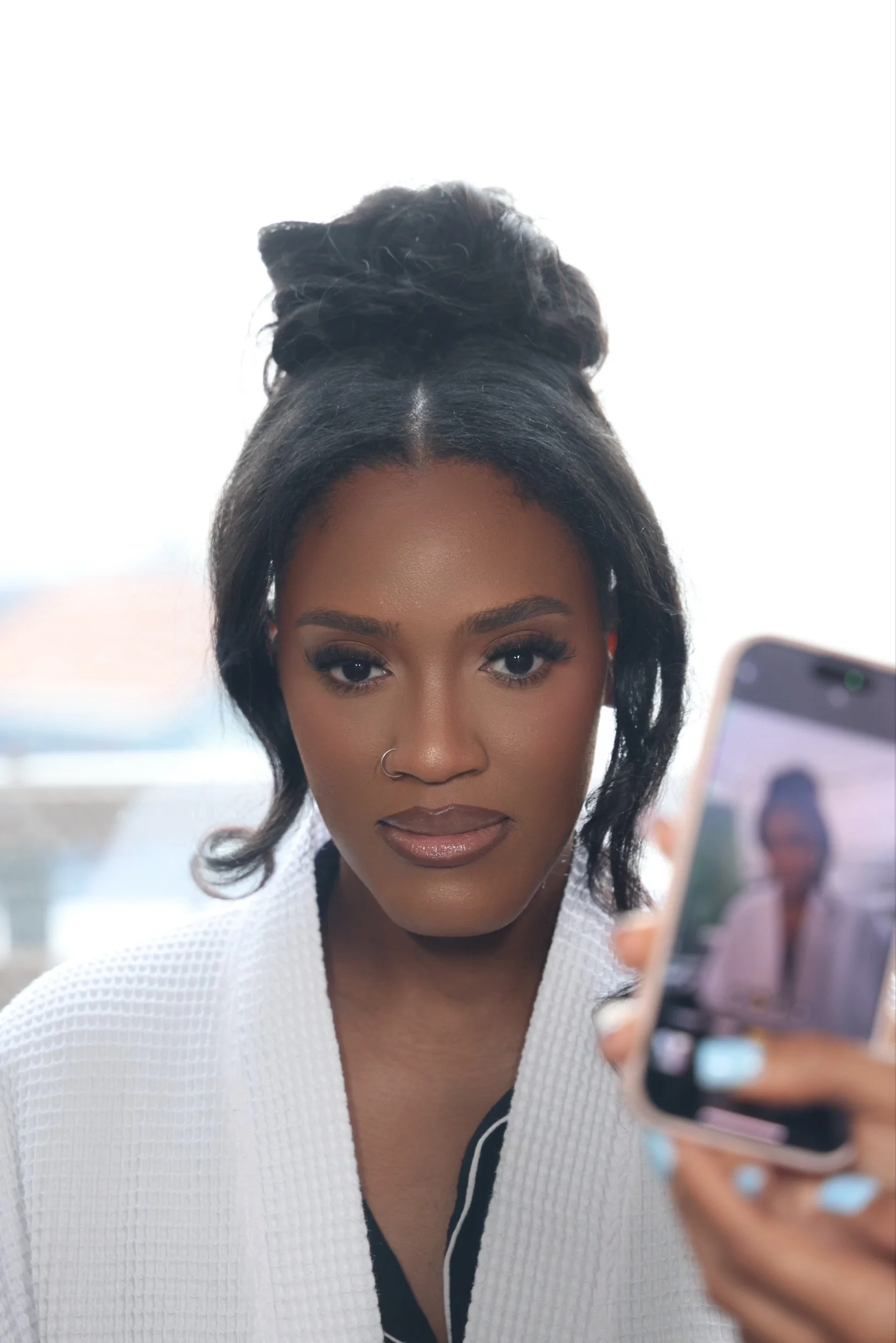 Close-up of a woman with dark skin, makeup, and hoop nose ring, holding a smartphone taking a selfie indoors with blurred background screens.